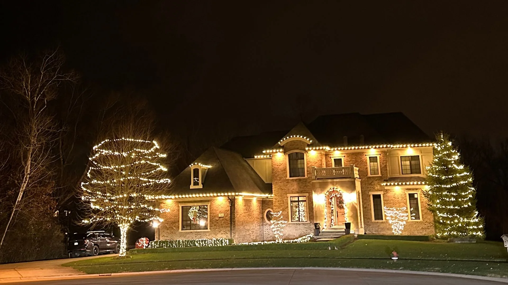 A large brick house decorated with Christmas lights at night. The house has a front porch, multiple windows, and nearby trees wrapped in string lights. The yard is well-maintained with a small lawn and bushes outlined with lights.