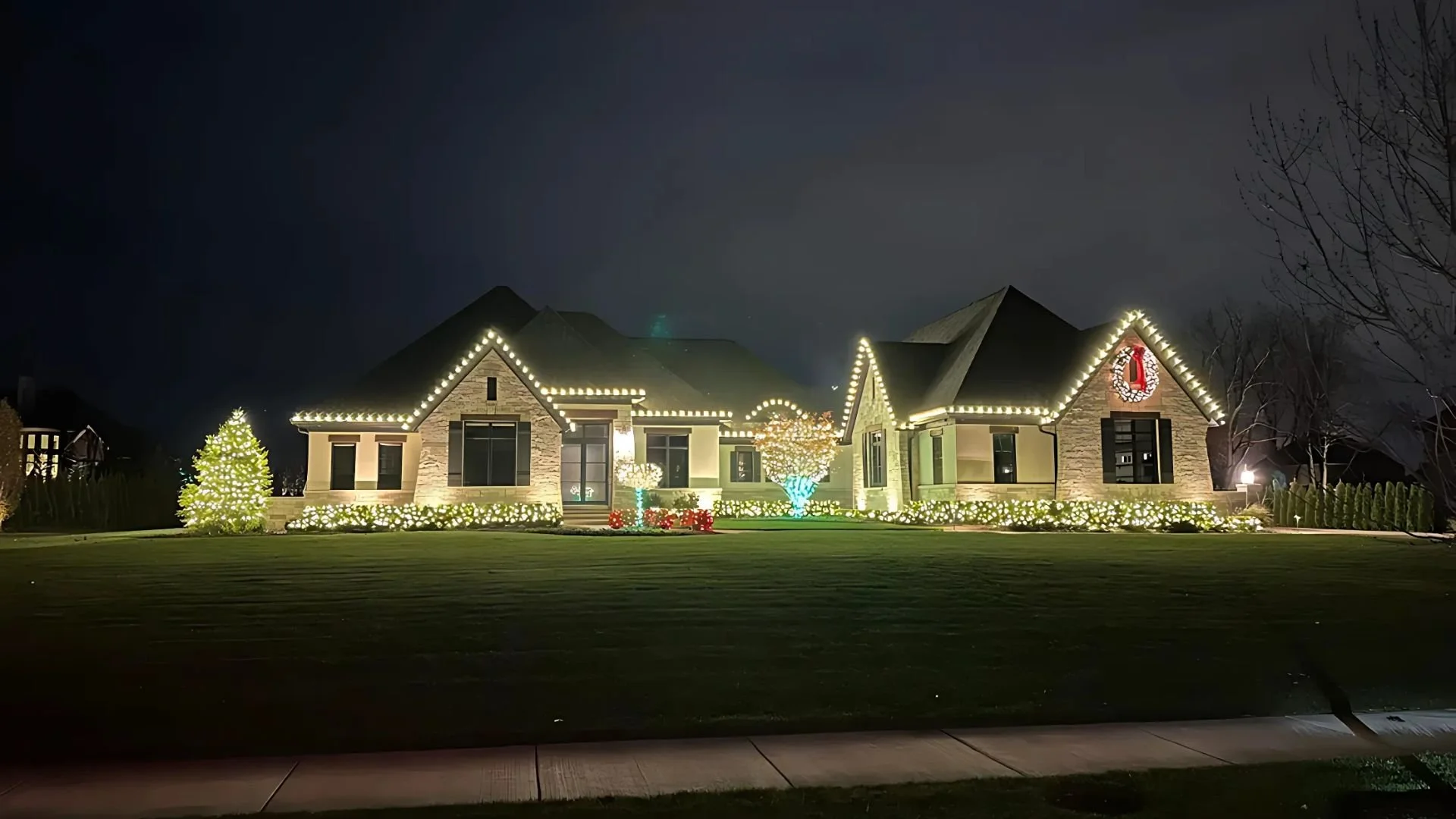 A house decorated with Christmas lights at night, featuring lit-up windows, a Christmas wreath, and Christmas trees in the front yard.