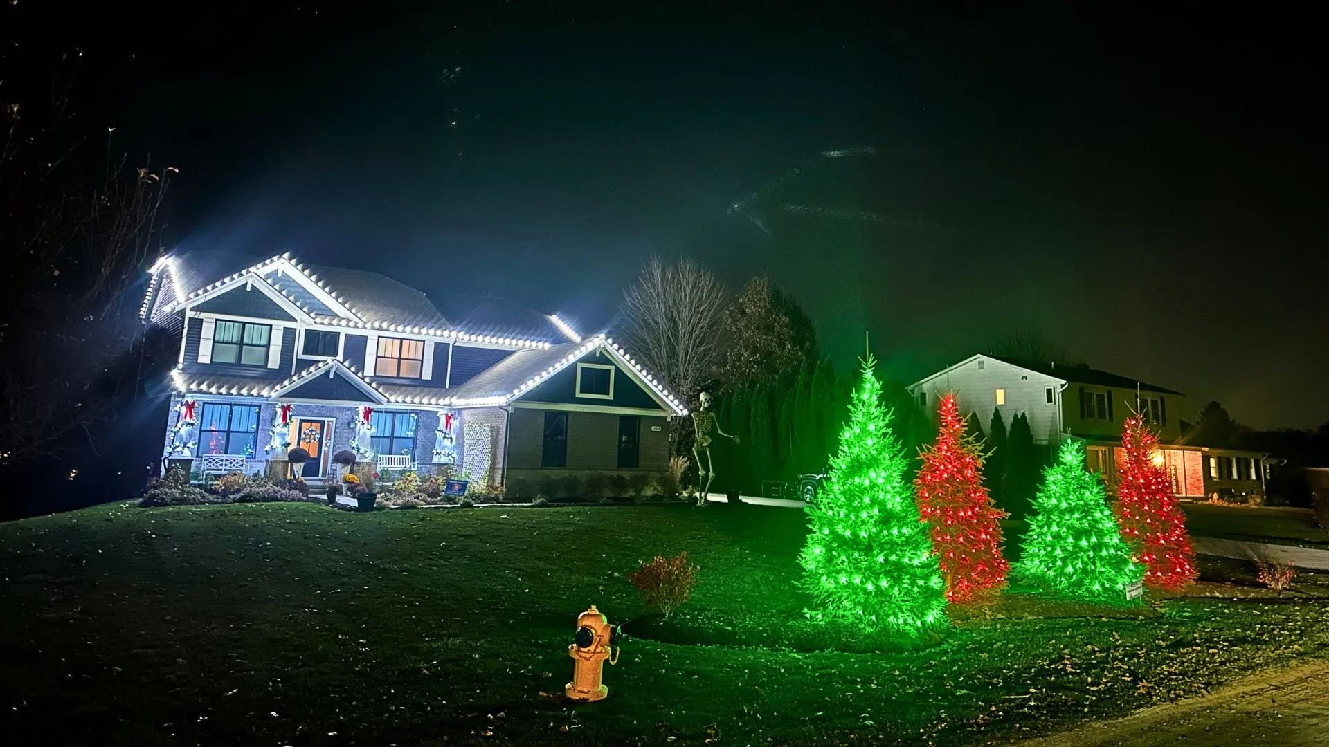 Nighttime holiday light display on a two-story house with white trim and roof decorated with white lights. In front, there are two green Christmas trees and two red Christmas trees illuminated with lights, and a small yellow fire hydrant on the lawn.