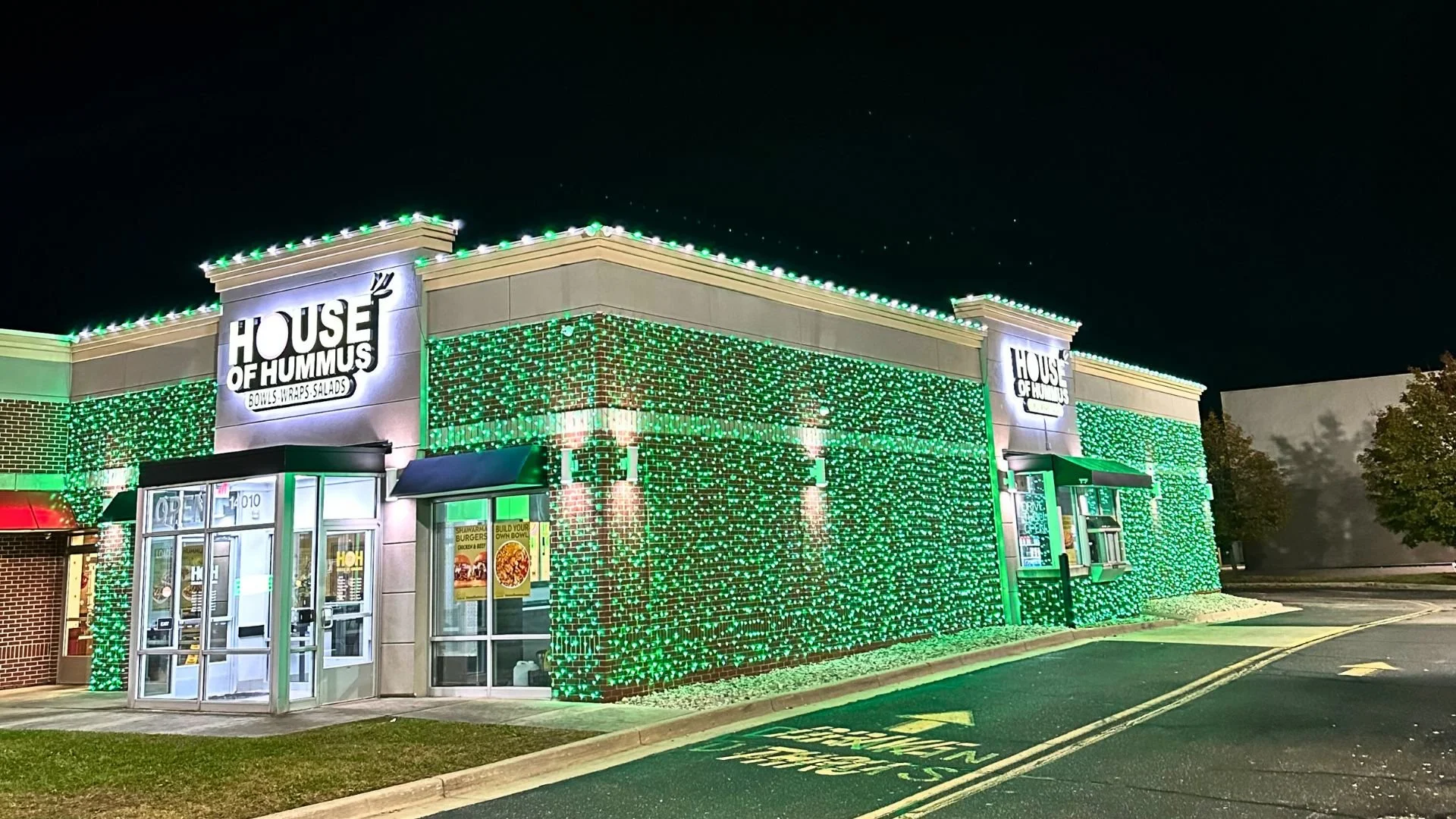 Night view of a fast-food restaurant called 'House of Hummus' decorated with green holiday lights, with a drive-thru window and a sign advertising build your own bowl.