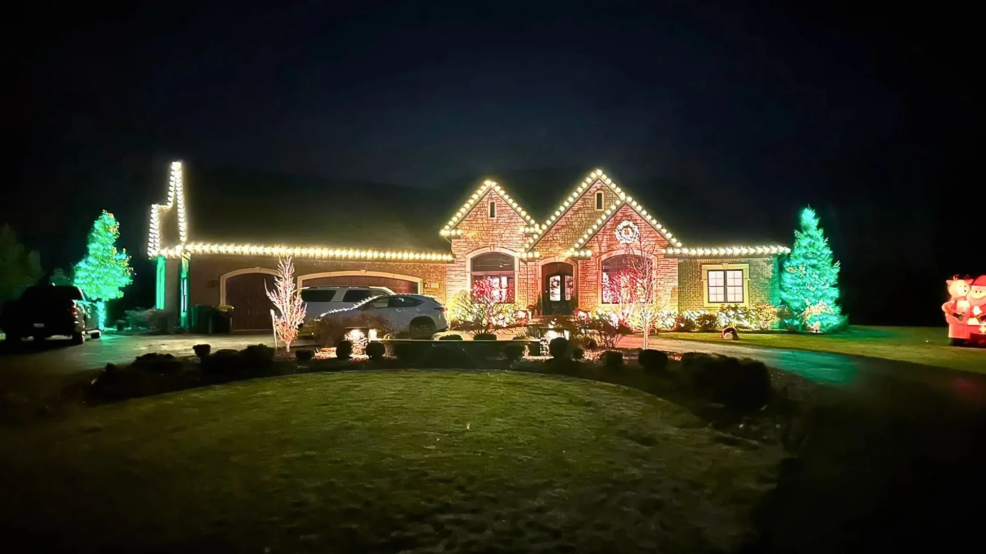 A house decorated with Christmas lights at night, featuring illuminated trees, bright window lights, and inflatable Christmas figures outside.