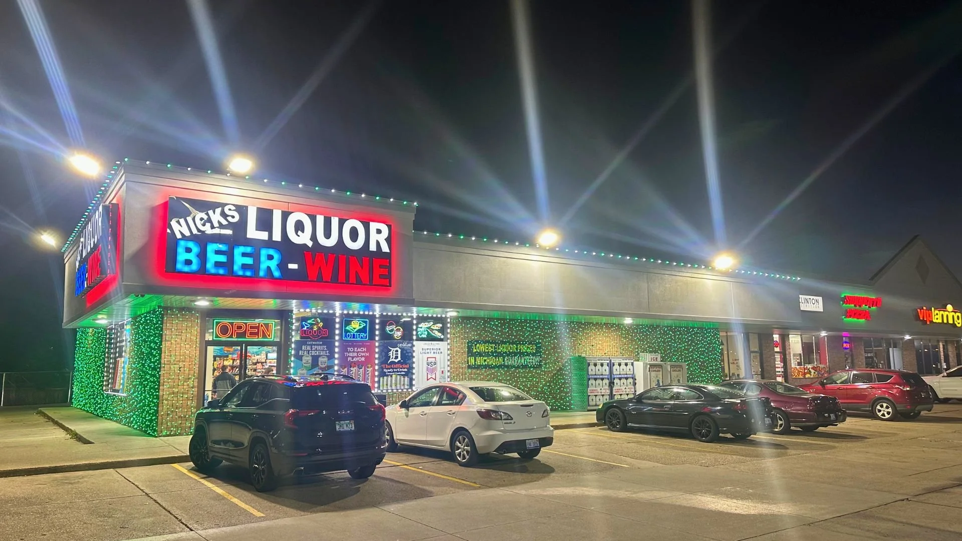 Night view of a liquor store with a bright neon sign advertising liquor, beer, and wine. The store's exterior is decorated with green lights, and there are several cars parked in front.