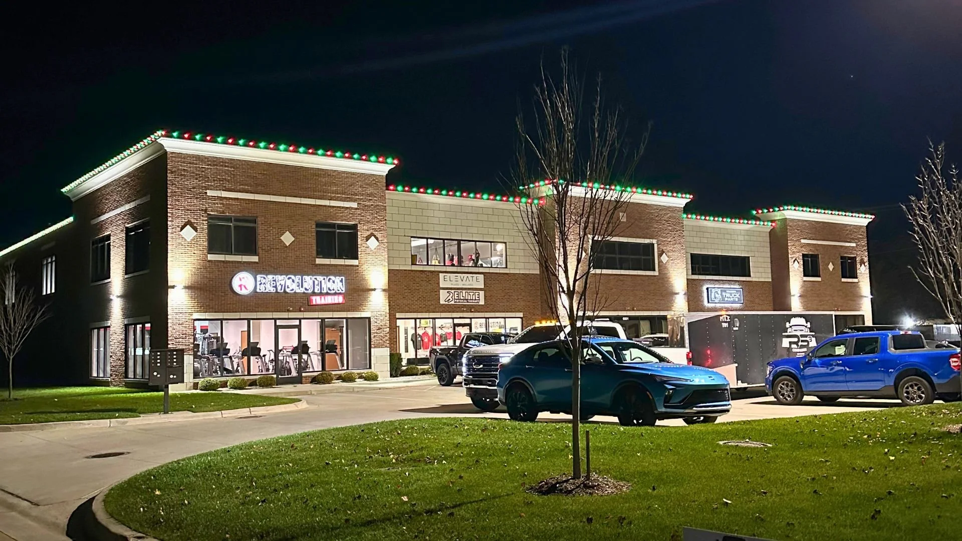 Night view of a two-story commercial building decorated with holiday lights on the roofline. The building has brick exteriors with large windows showing fitness equipment inside. There are several parked cars in front, including a blue SUV, a black s