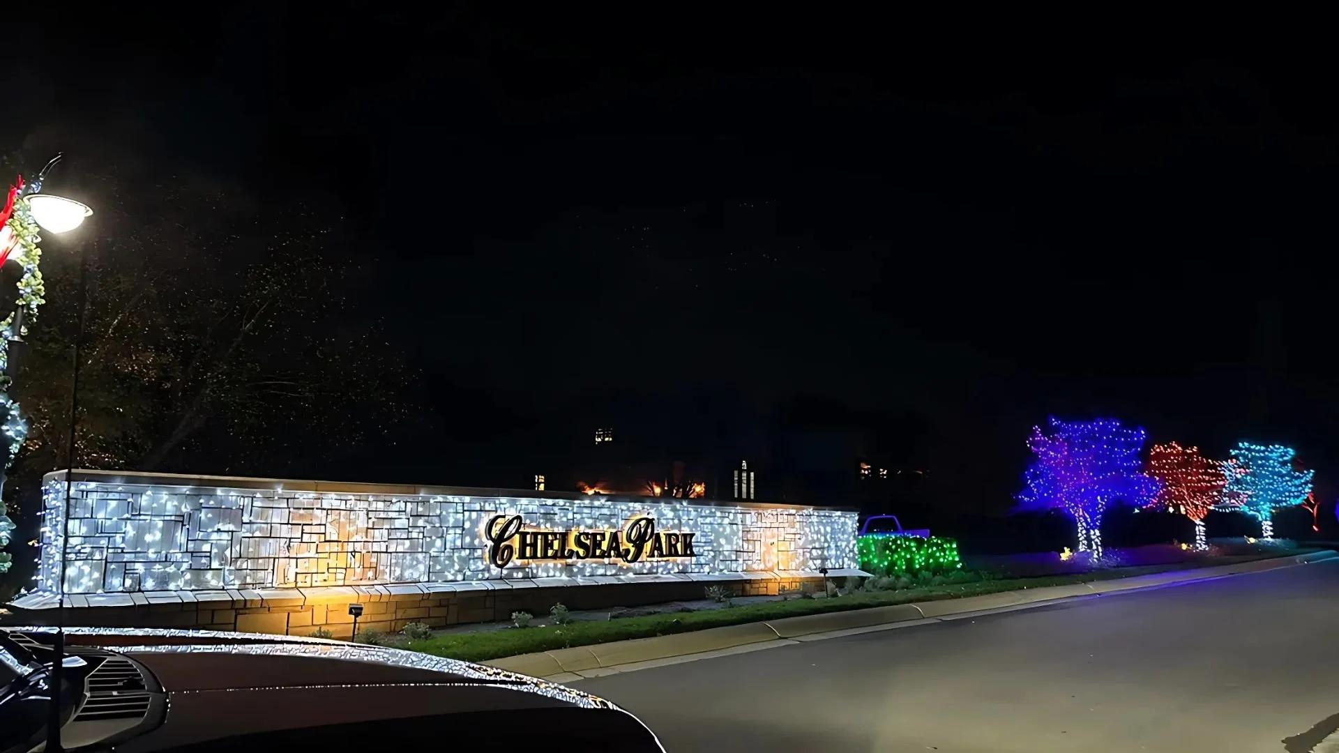 Night view of Chelsea Park entrance with festive holiday lights, illuminated trees in blue, red, and purple, and a decorated sign reading 'Chelsea Park' on a brick wall.