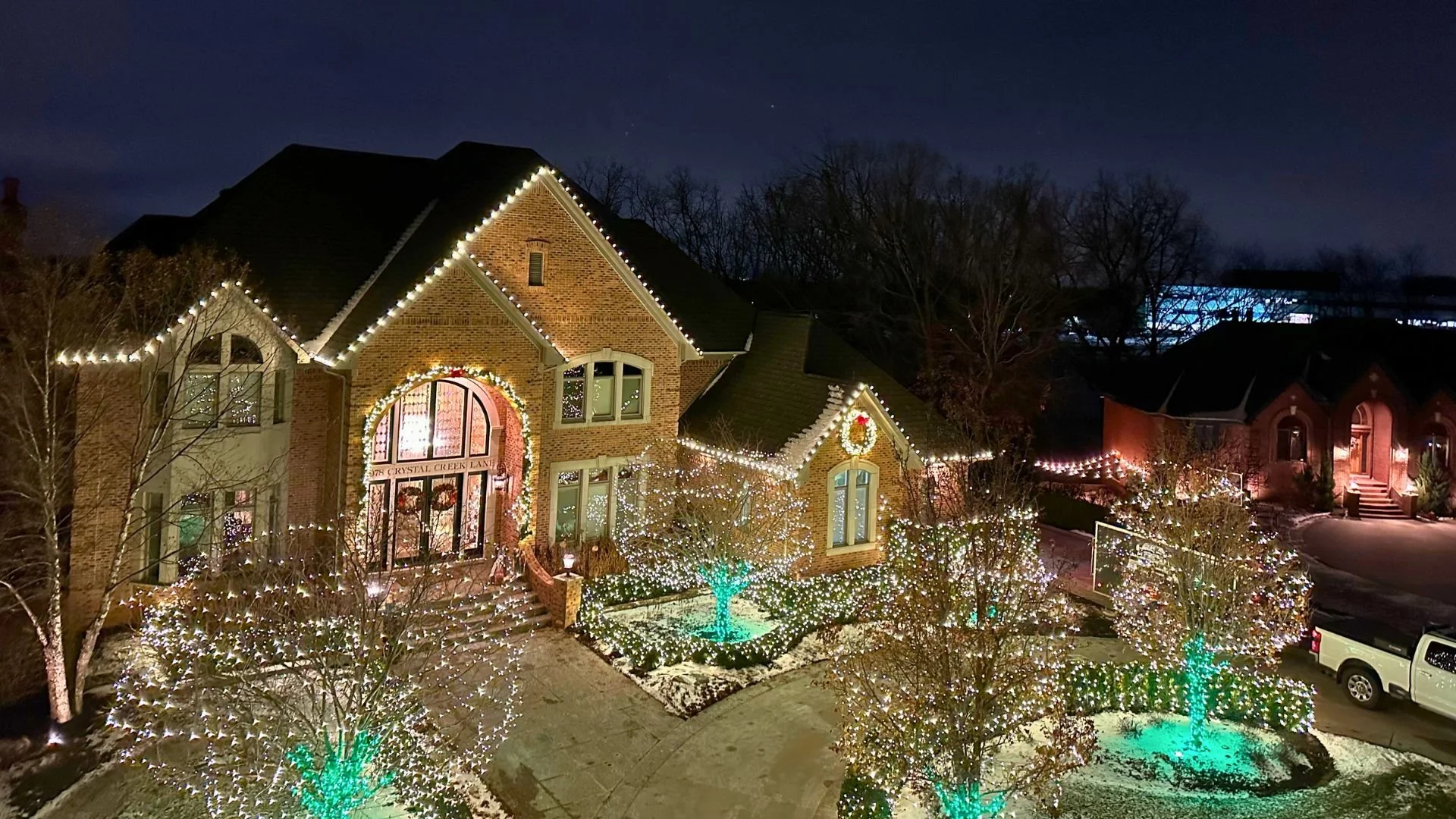 A large brick house decorated with Christmas lights and wreaths at night, with snow on the ground, illuminated trees, and neighboring houses.