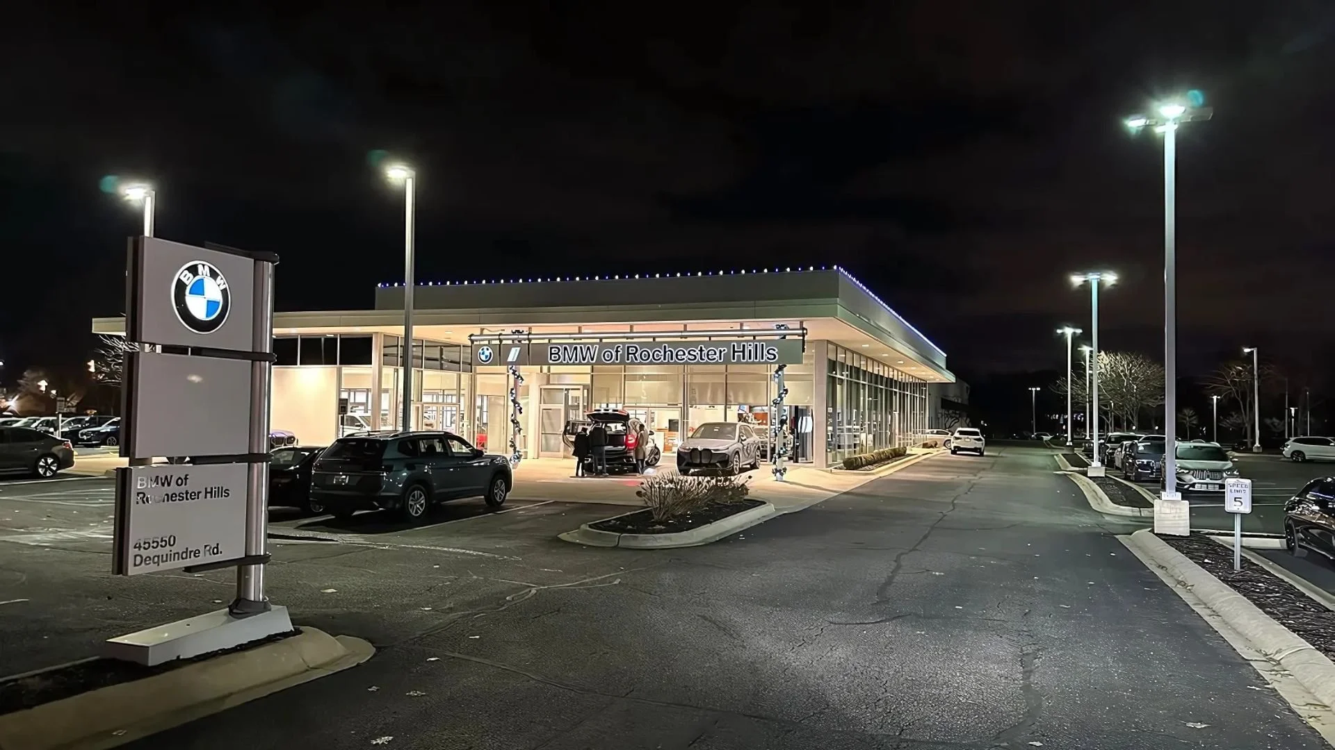 Night view of BMW dealership in Rochester Hills with multiple cars parked outside, illuminated by tall lights, featuring a modern building with a glass facade and multiple cars inside and outside.