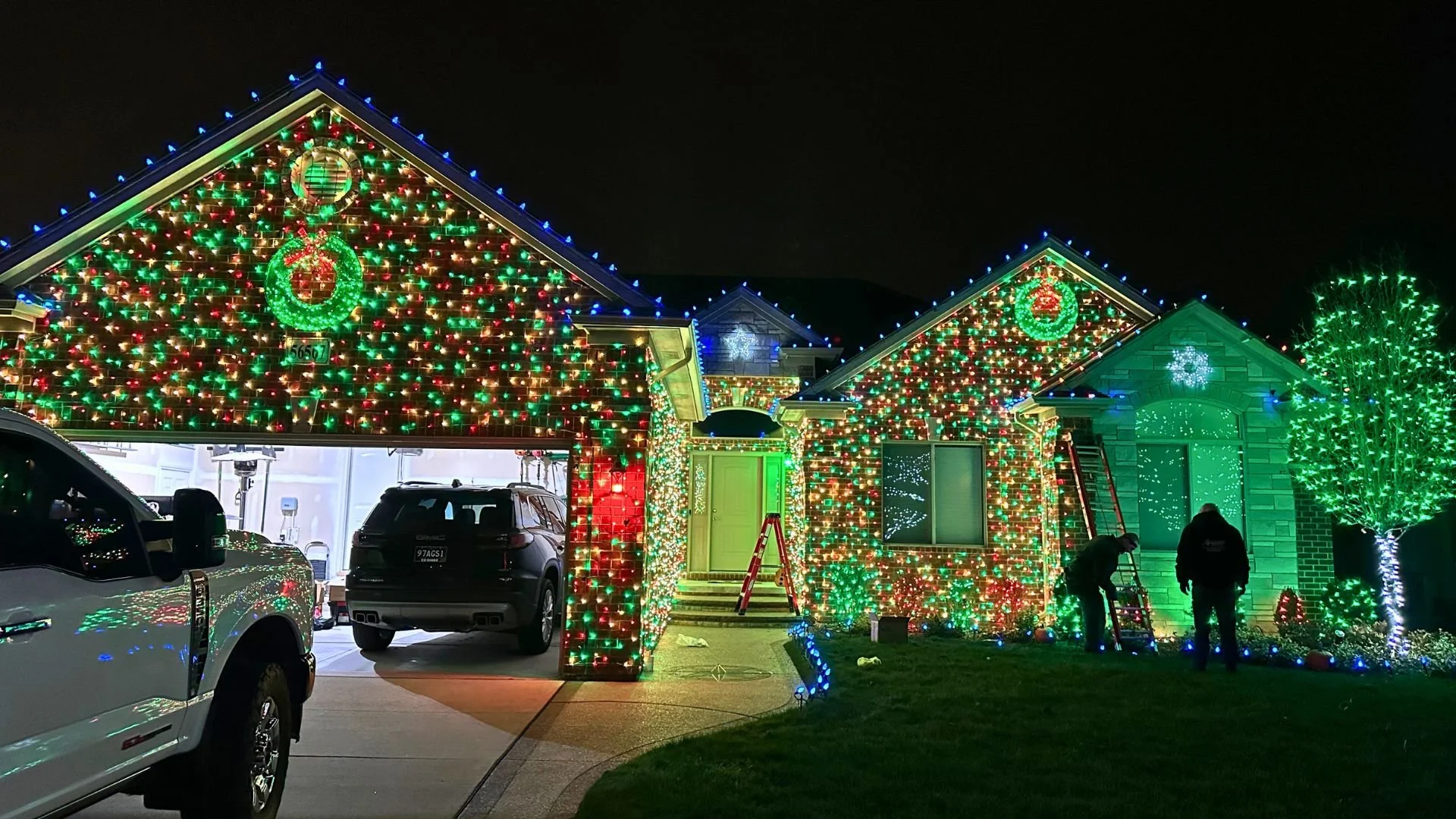 House decorated with colorful Christmas lights, wreaths, and ornaments at night, with cars parked in the driveway and people working on the decorations.