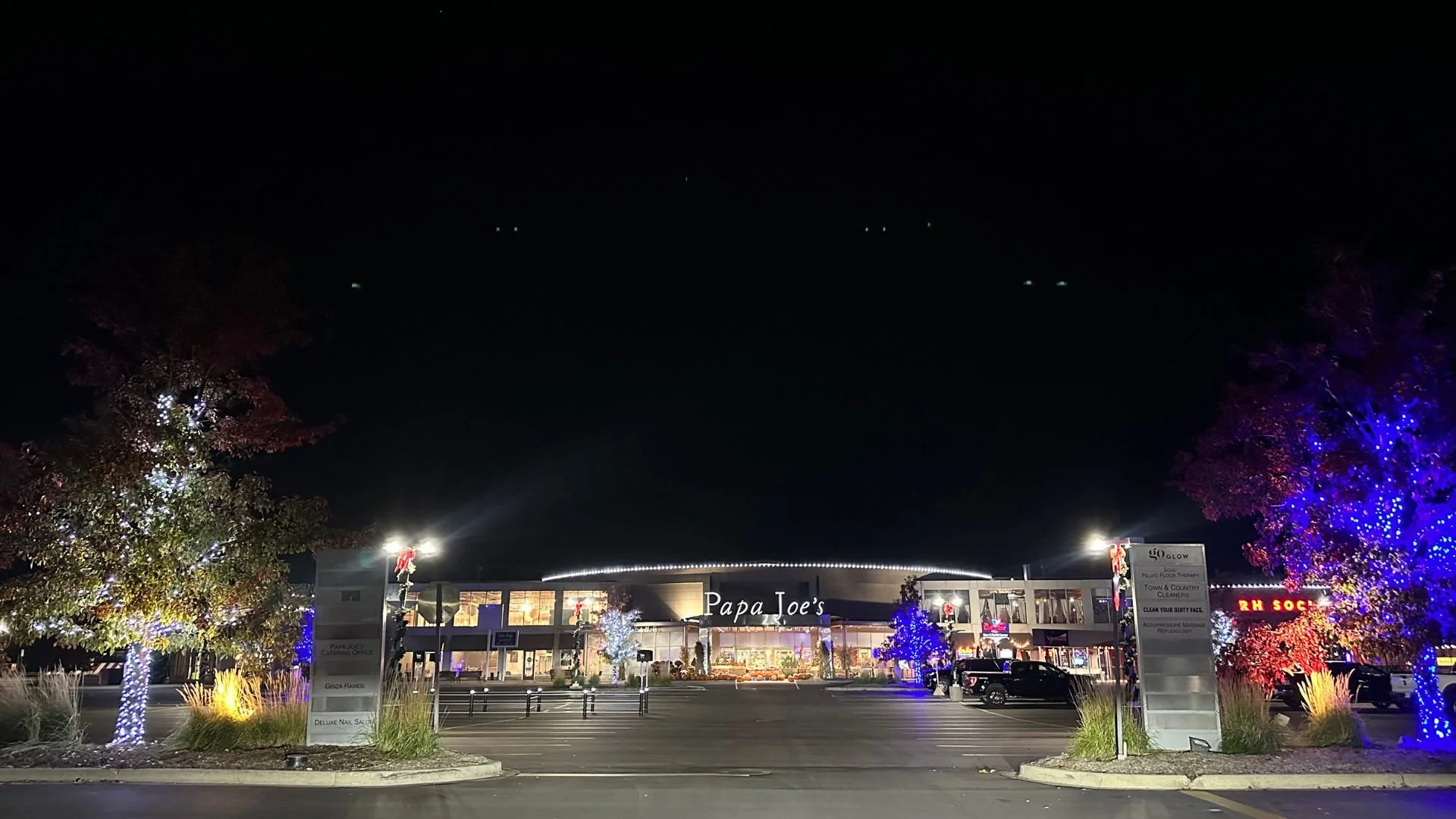Night view of a shopping plaza with colorful lit trees, a Papa Joe's store in the center, and parking lot in front.