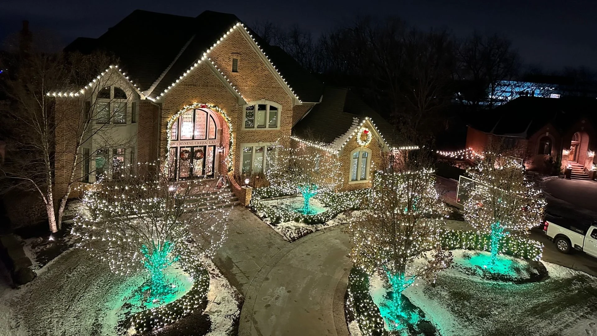 A large brick house decorated with Christmas lights and wreaths, with trees illuminated by turquoise lights in the yard, at night during winter.