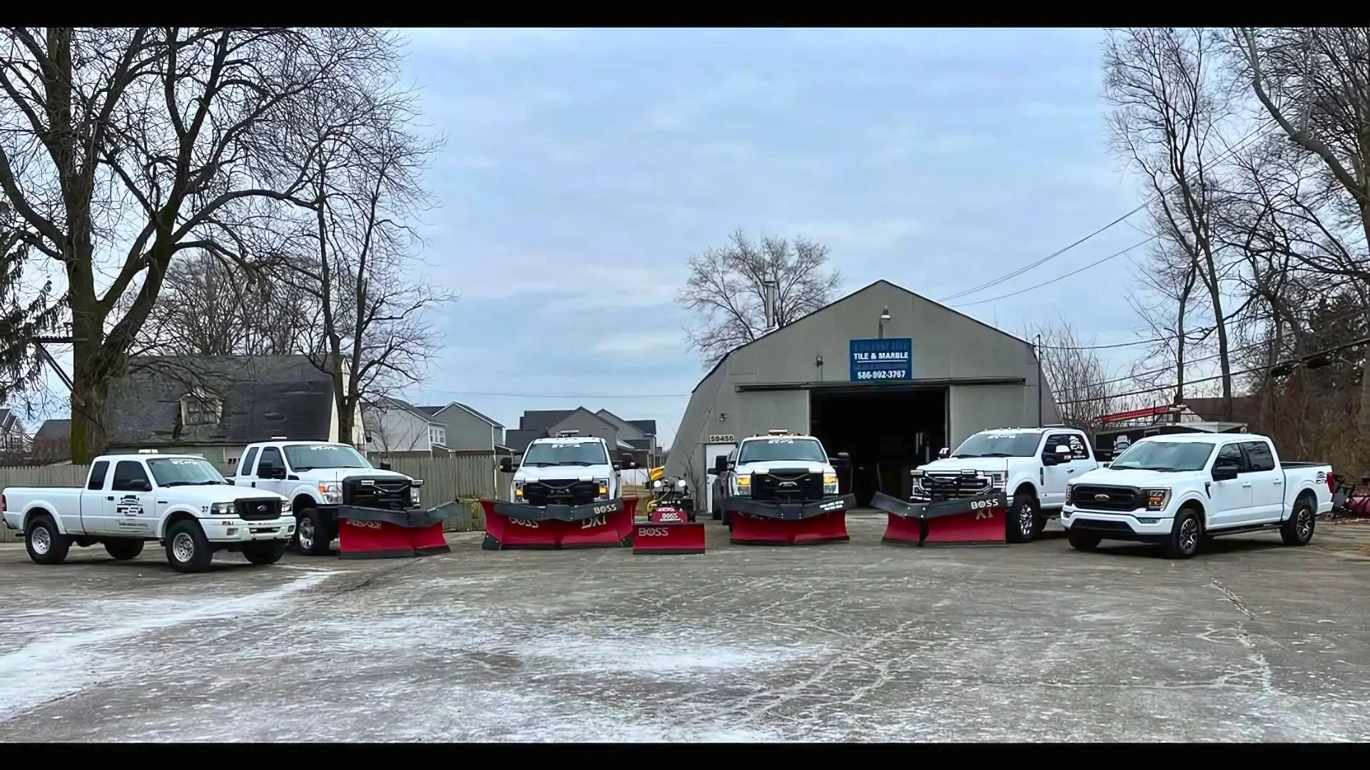 A row of six pickup trucks parked outside a large gray metal building with a blue sign that reads 'Tile & Marble'. The trucks are equipped with snow plows and are white with black and red accents. The surrounding area has a few leafless trees, a wood