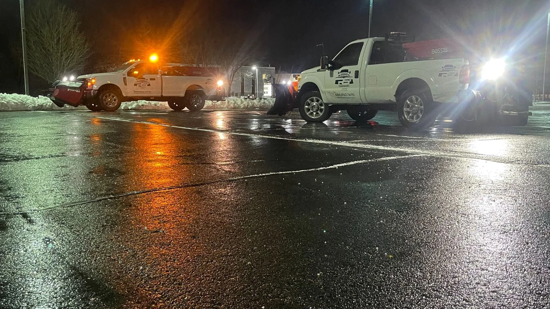 Nighttime scene in a wet parking lot with two police or emergency vehicles, one white pickup truck and one larger SUV, parked with their lights flashing. Snow piles are visible along the edges of the parking lot.
