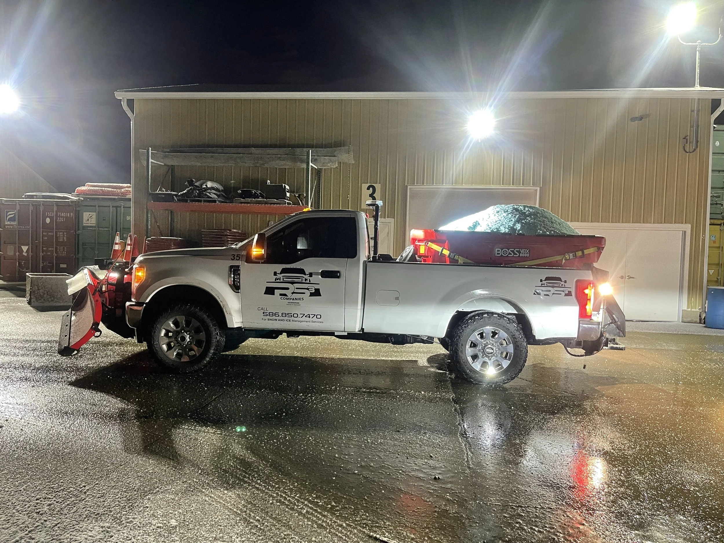 White utility vehicle with snow plow attached, and snowmobile in the truck bed, parked outside a building at night with bright security lights