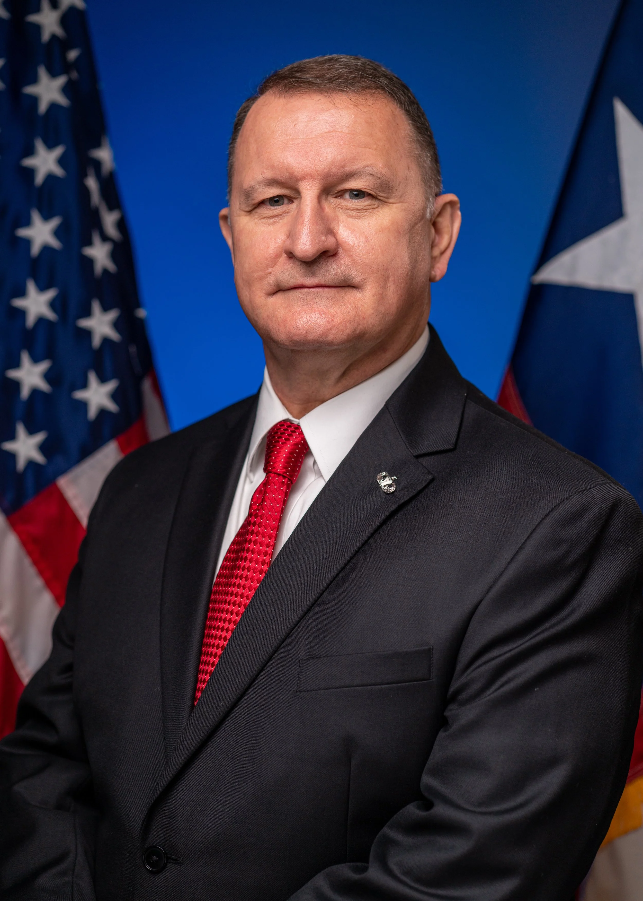 A man in a dark suit, white shirt, red patterned tie, standing with arms crossed, in front of American and Texas flags.