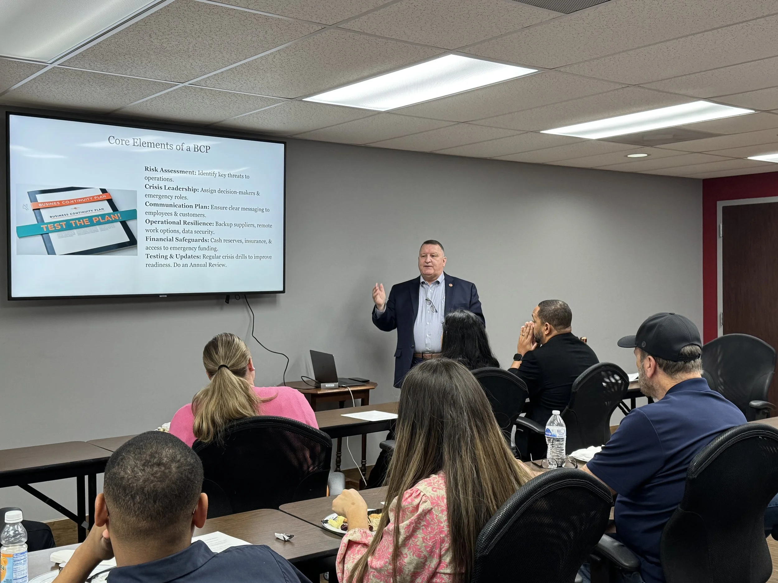 A man in a suit giving a presentation in a conference room with people seated at tables, watching and listening.