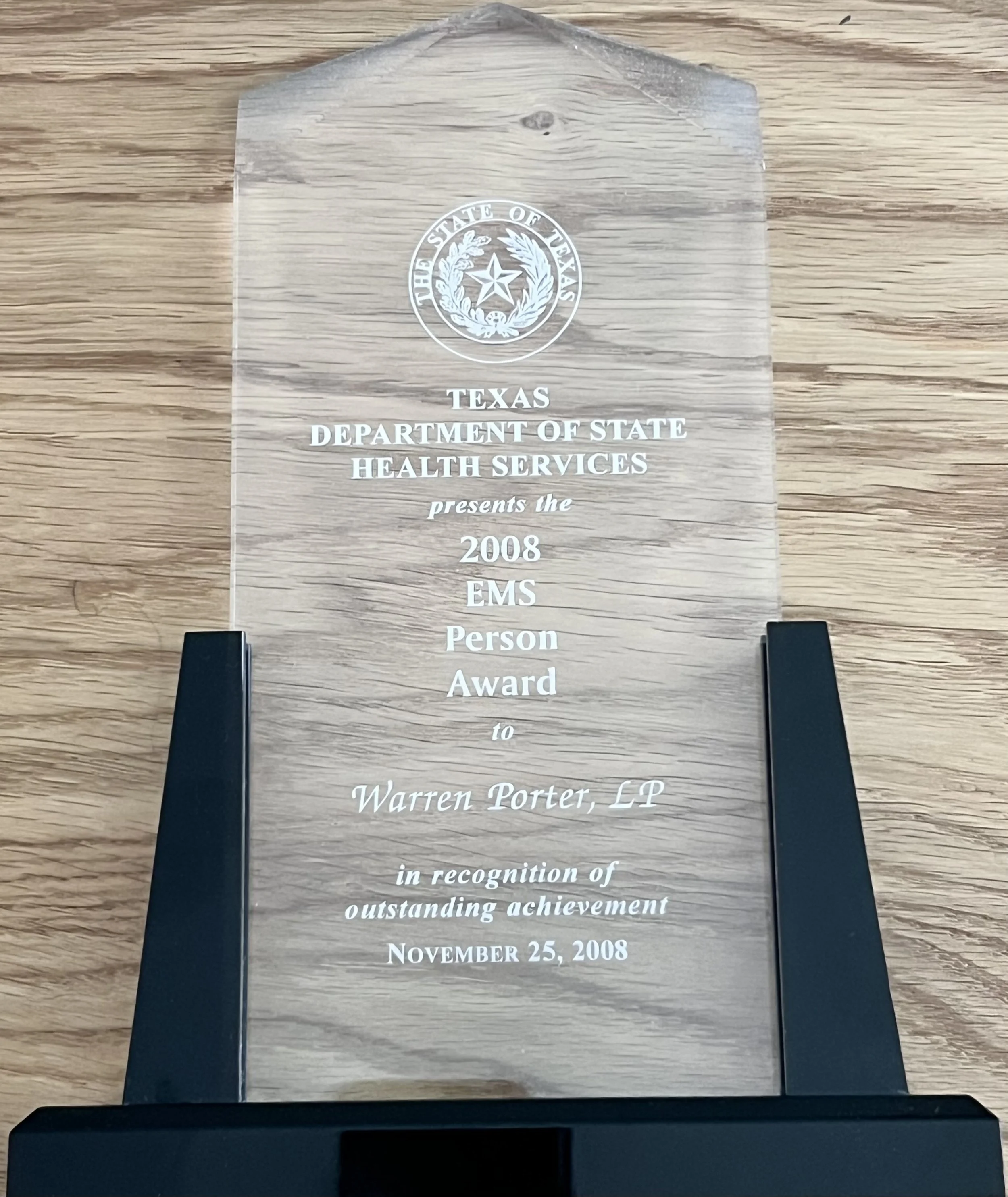 A clear, engraved glass award with a wooden background. The text on the award reads: 'The State of Texas, Texas Department of State Health Services presents the 2008 EMS Person Award to Warren Porter, LP in recognition of outstanding achievement, November 25, 2008.'