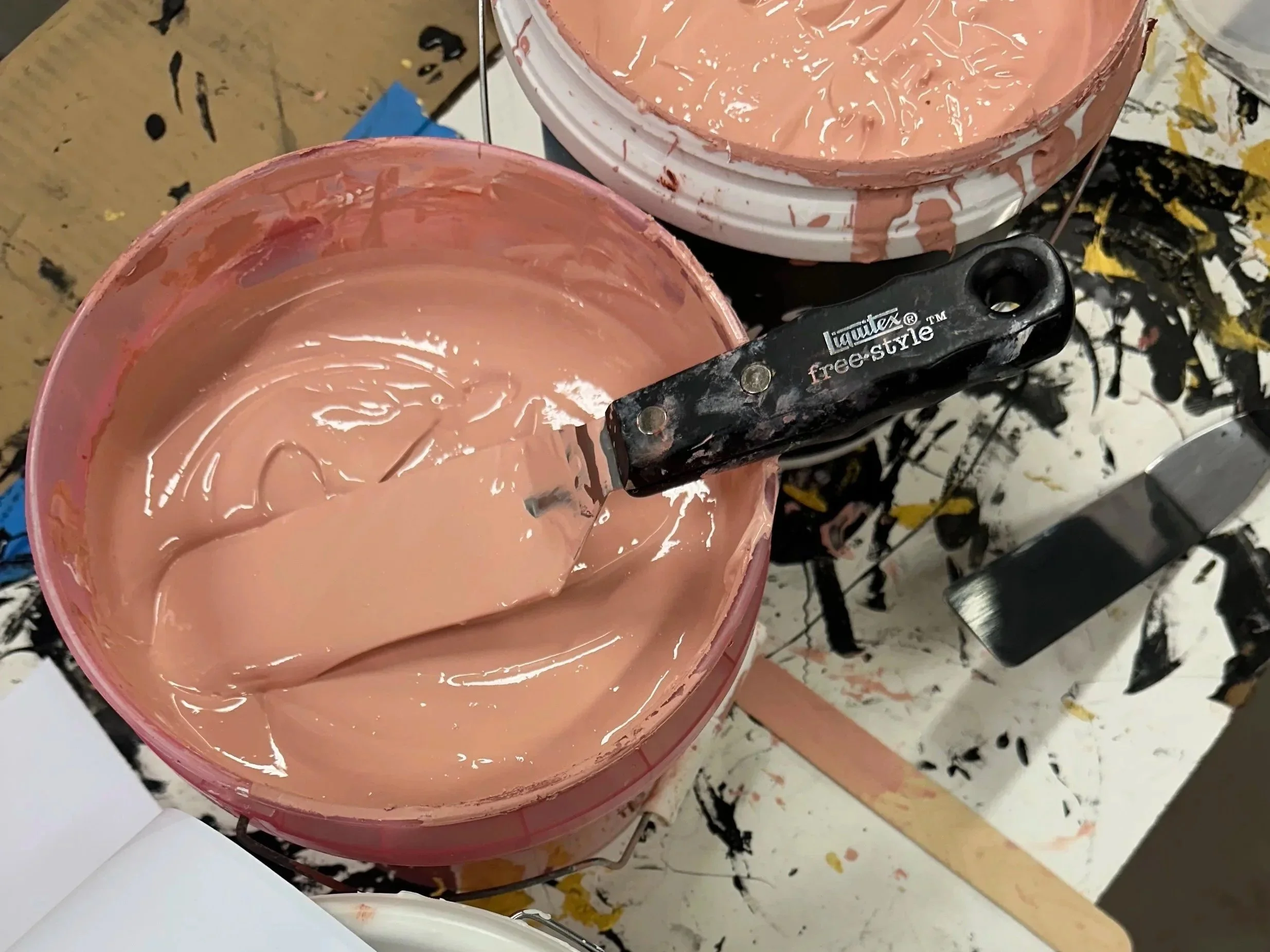 A close-up view of two open buckets filled with pink paint, with a paint stirrer resting on one of the buckets, on a table covered with splatters of black, brown, and pink paint.