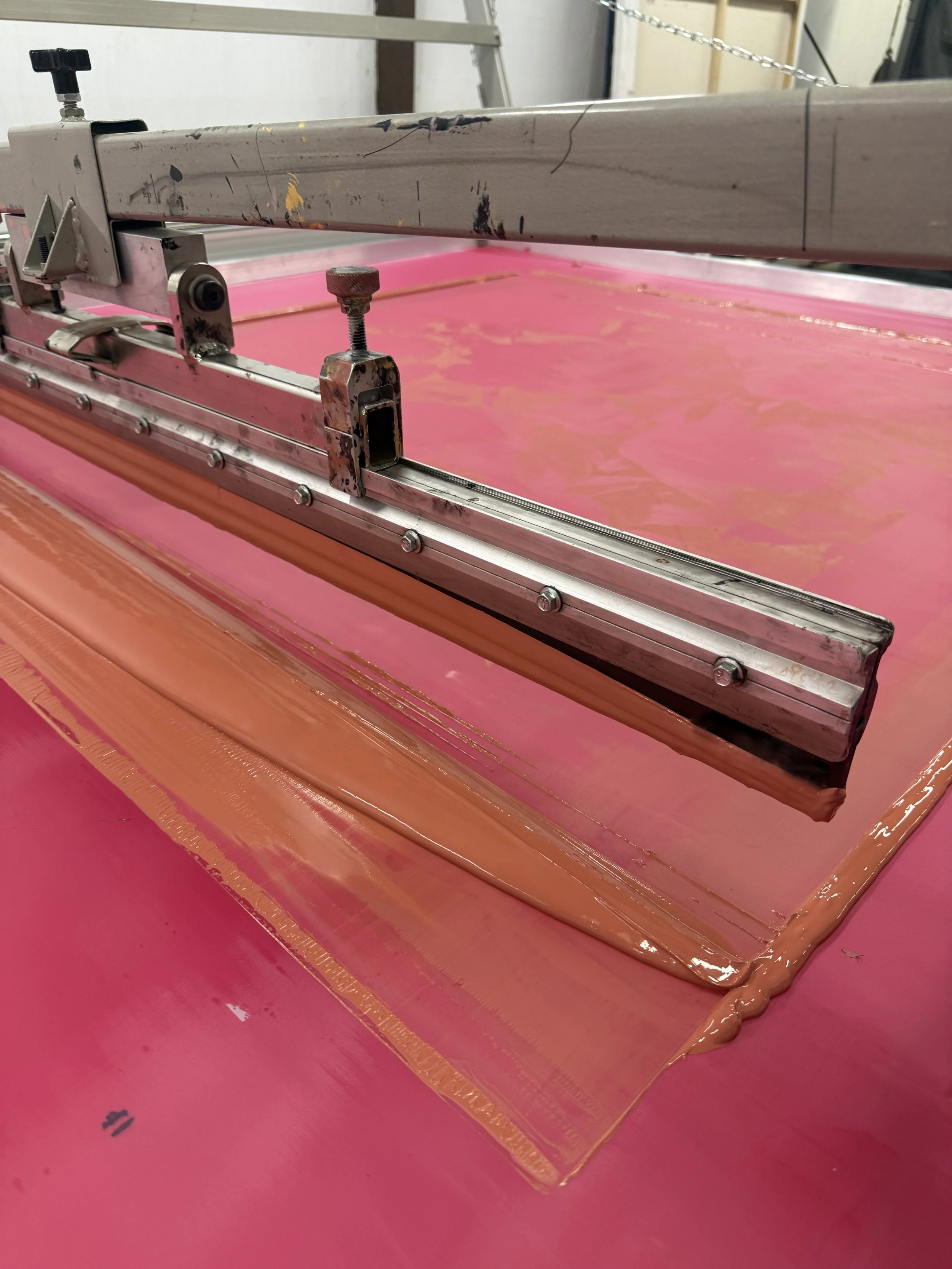 Close-up of a printing or dyeing table with pink fabric and liquid dye, with metal rail and clamps.