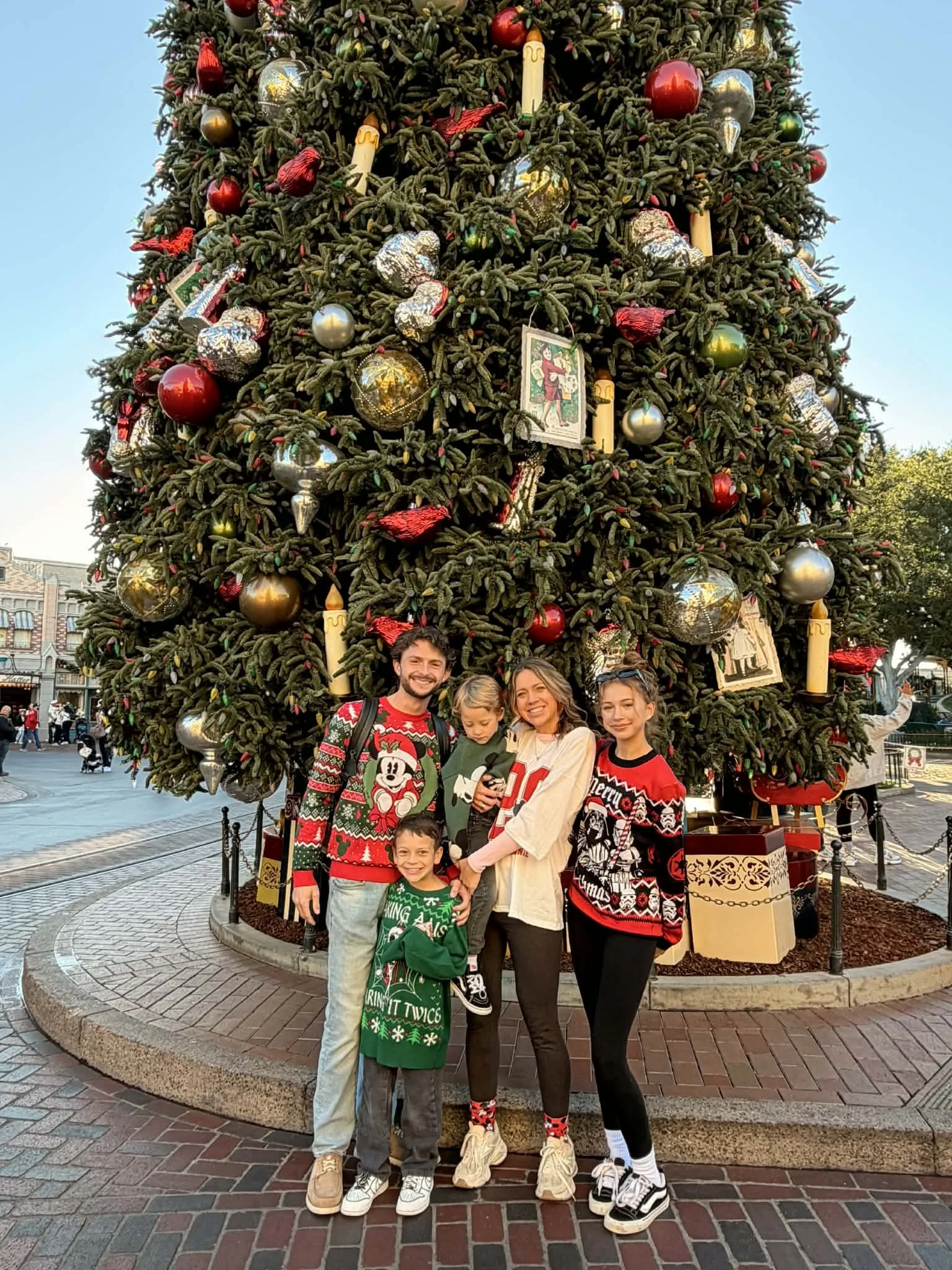 A family of five standing in front of a large decorated Christmas tree at an outdoor location, wearing festive holiday sweaters and smiling.