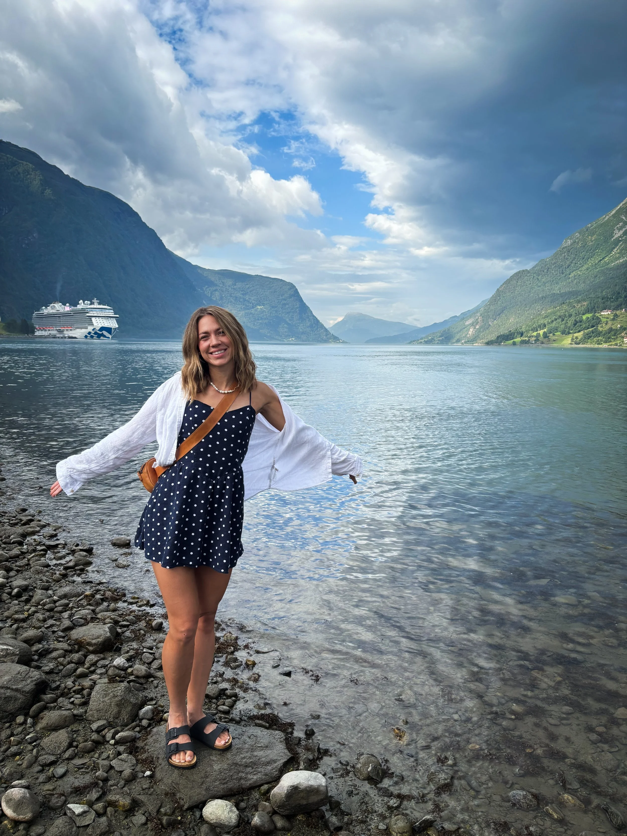 A smiling woman in a navy polka dot dress and white cardigan standing on a rocky shoreline by a lake, with mountains and a cruise ship visible in the background under a partly cloudy sky.