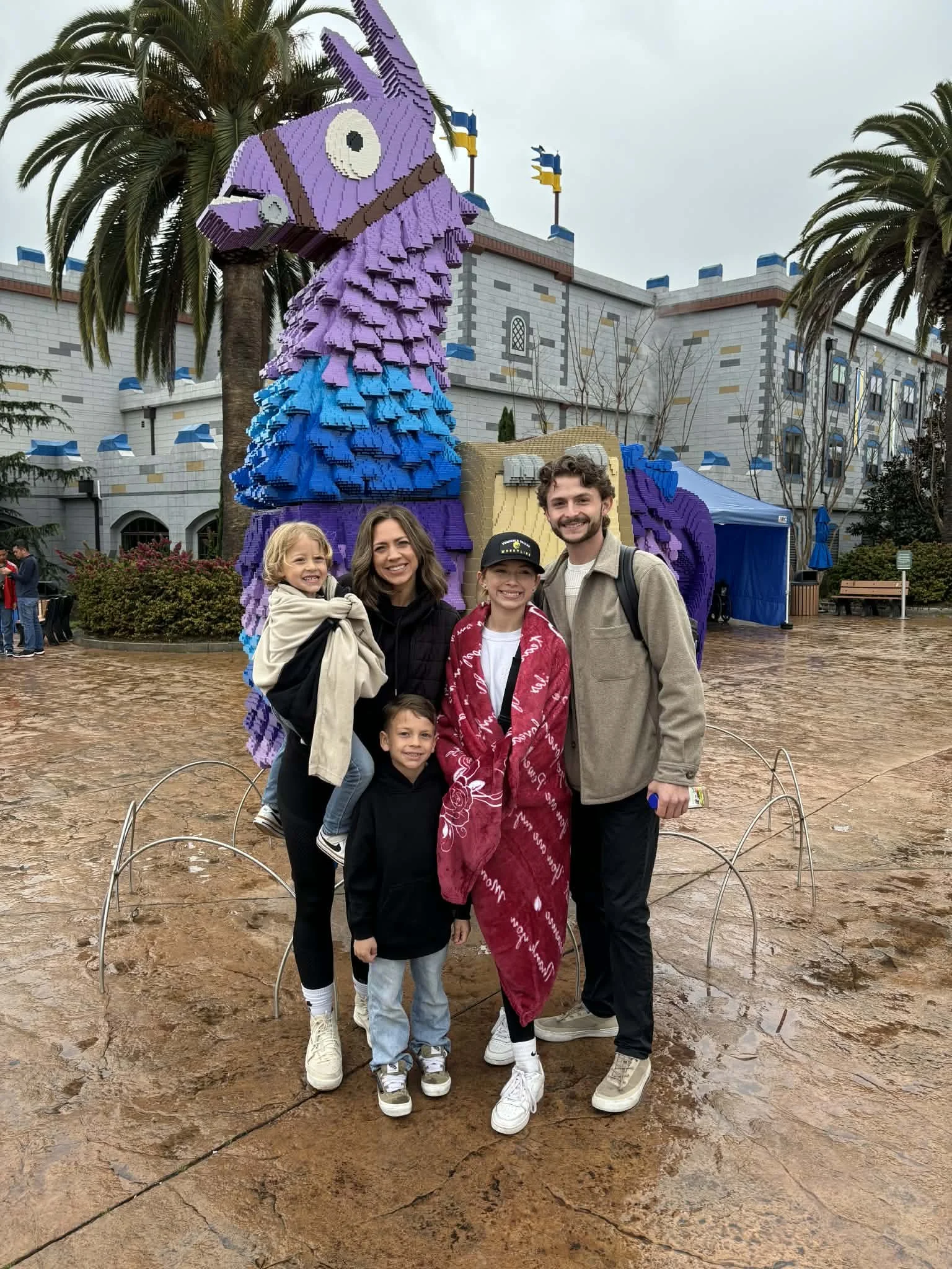 A family of five standing in front of a large, colorful llama sculpture made of LEGO bricks at a theme park. The sculpture is purple, blue, and brown, and is set outdoors with palm trees and a castle-like building in the background. The family members are smiling and wearing casual clothing, with some holding onto each other.