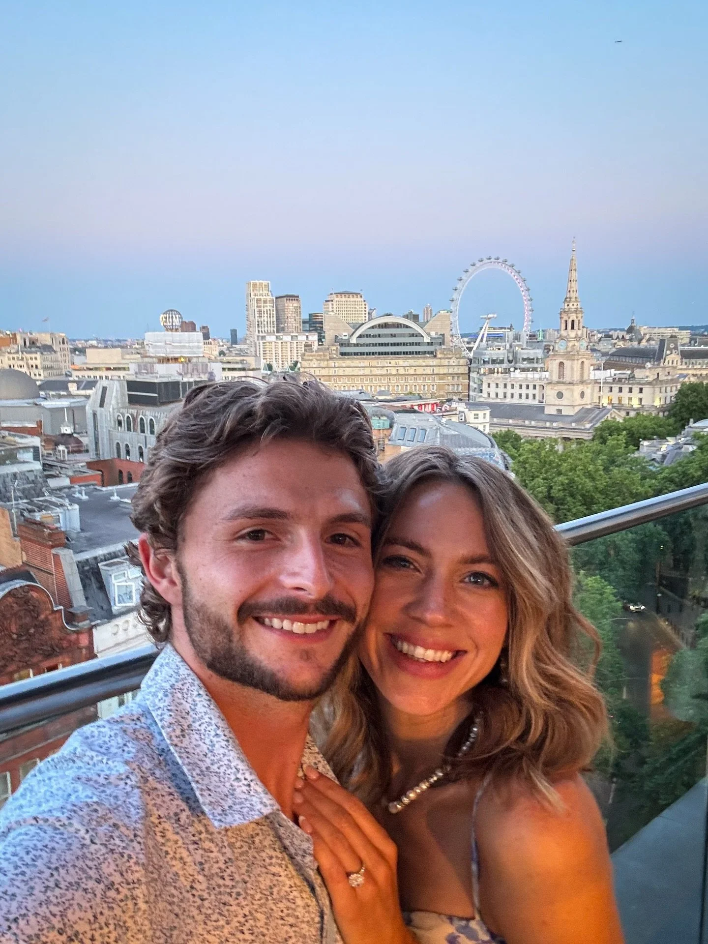 A smiling couple taking a selfie on a balcony with a city skyline in the background, including the London Eye and a church spire, during twilight.
