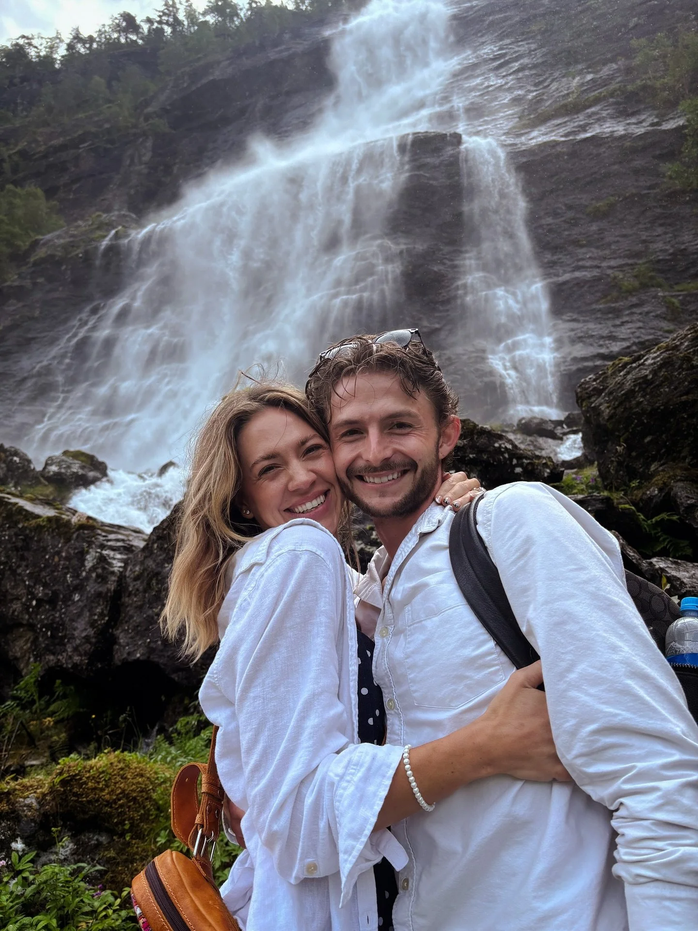A smiling couple hugging in front of a waterfall with rocks and greenery.