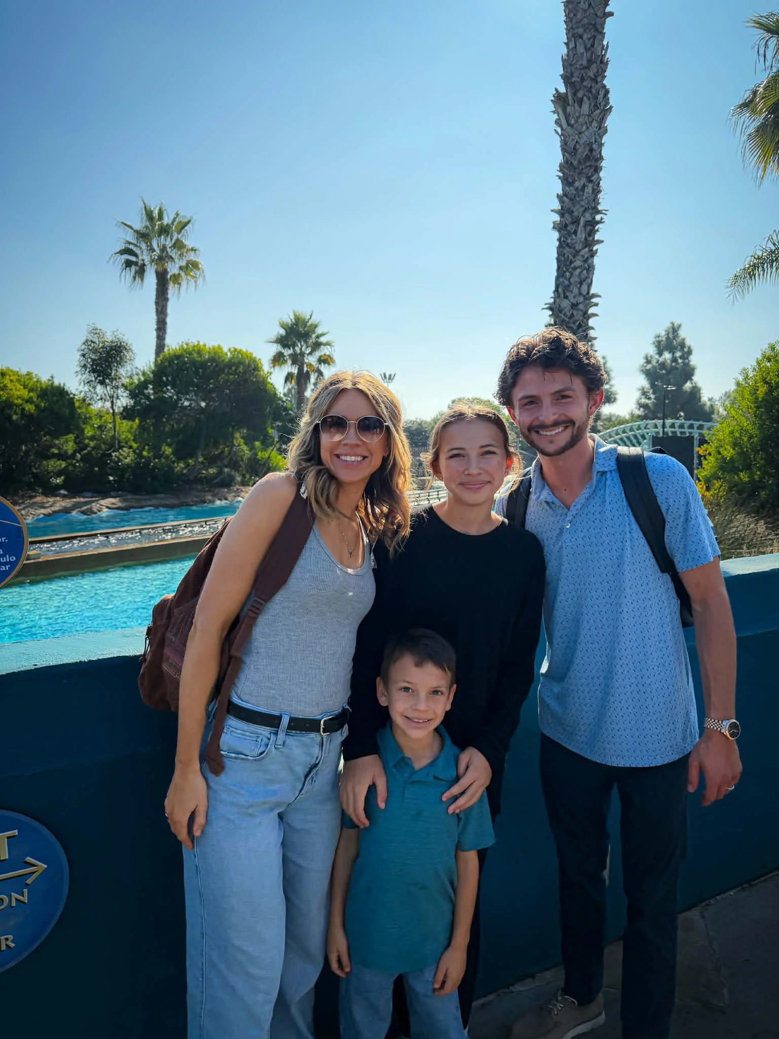 A family of four smiling, standing outdoors near a water feature with palm trees and greenery in the background, under a clear blue sky.
