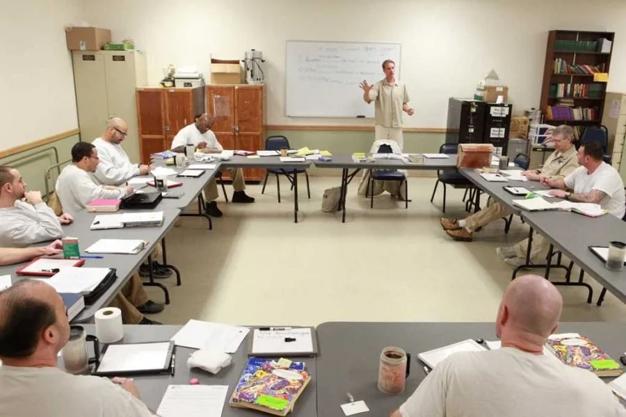 A classroom with a teacher standing in front of a whiteboard and students seated around tables arranged in a U-shape, engaged in a discussion or lecture.