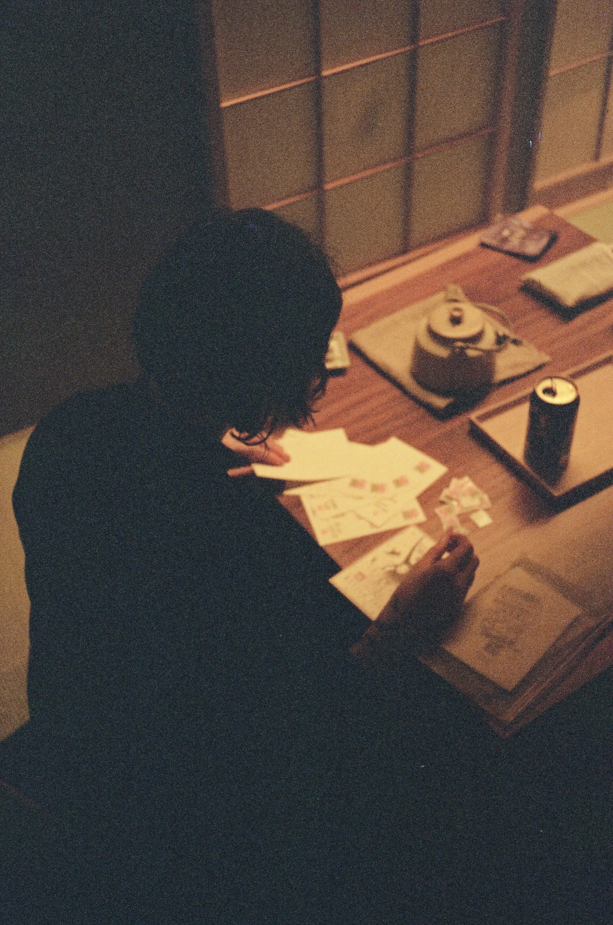 A person with dark hair is sitting at a wooden table, working on papers and drawings, with sake cups, a can of soda, and a disposable coffee cup on trays, in a traditionally styled room with shoji doors.
