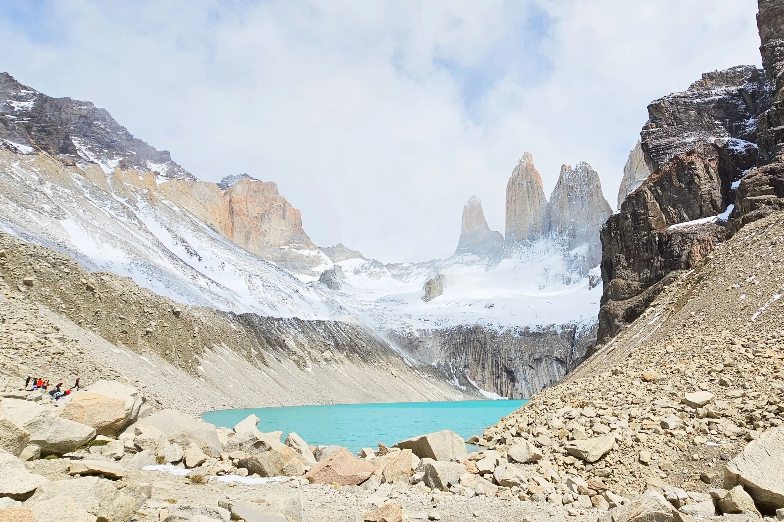 Snow-covered mountains surrounding a bright turquoise glacial lake with a few hikers along the rocky shoreline.
