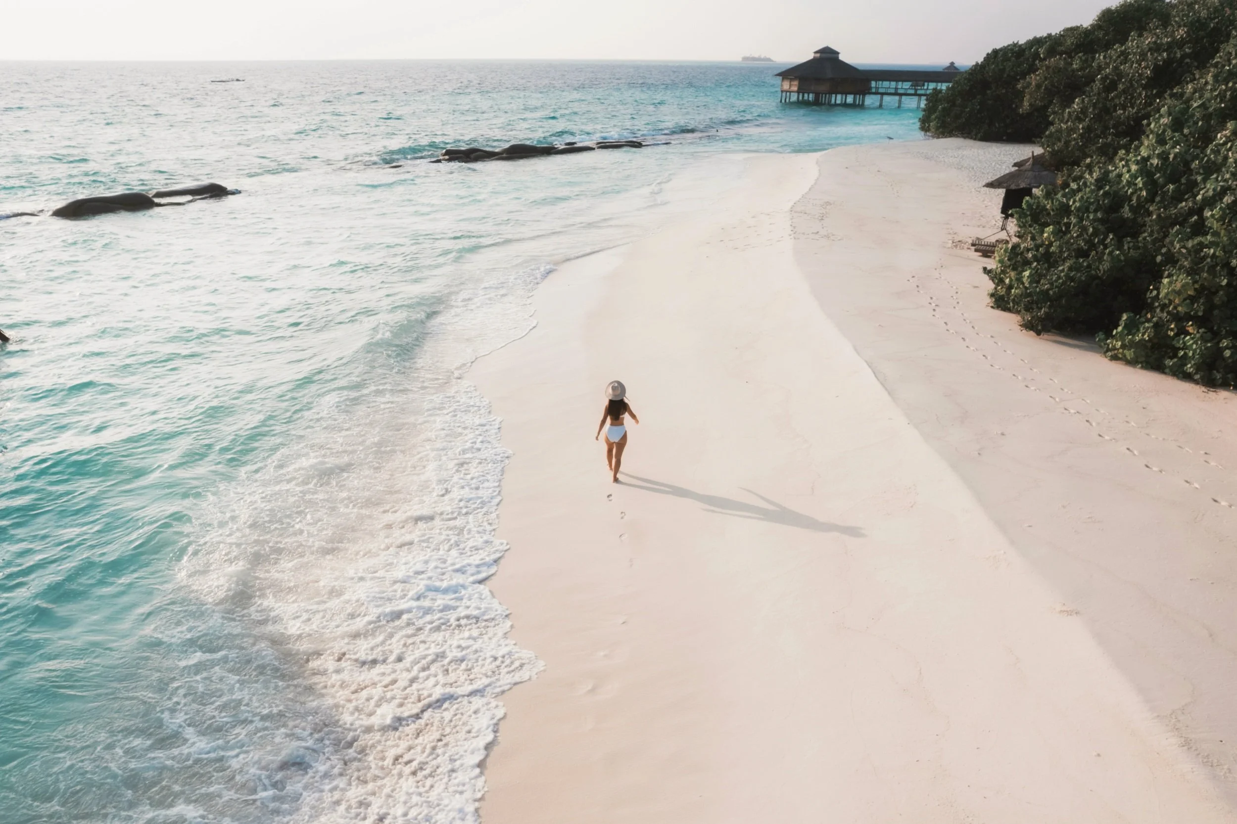 A woman walking alone on a white sandy beach near the ocean, with footprints behind her, during daytime with clear weather.