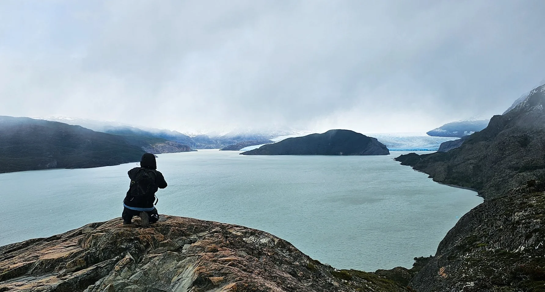 Hiker standing on a rock overlooking a blue-gray glacier lake surrounded by dark hills and a glacier in the distance