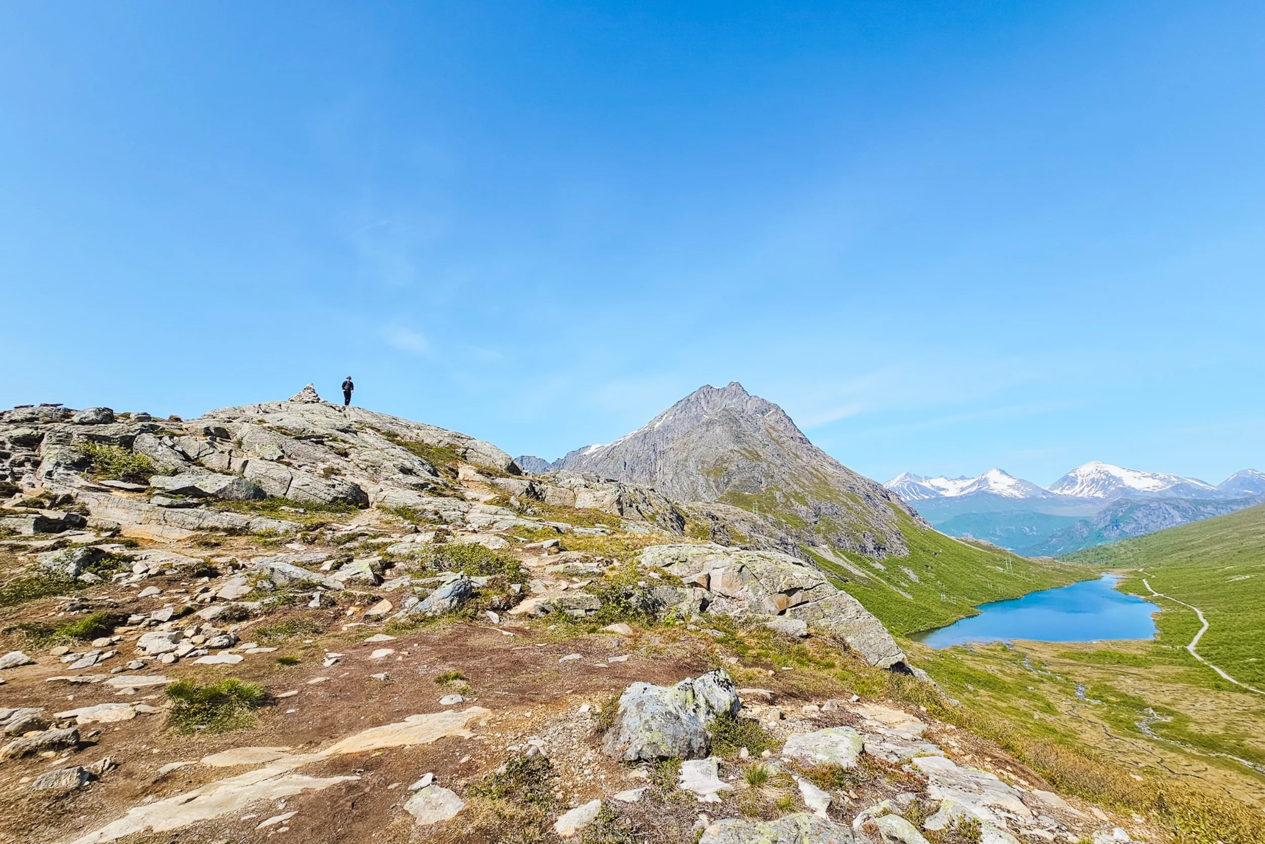 Man standing on rocks on a mountain overlooking a turquoise lake below and other snow capped peaks