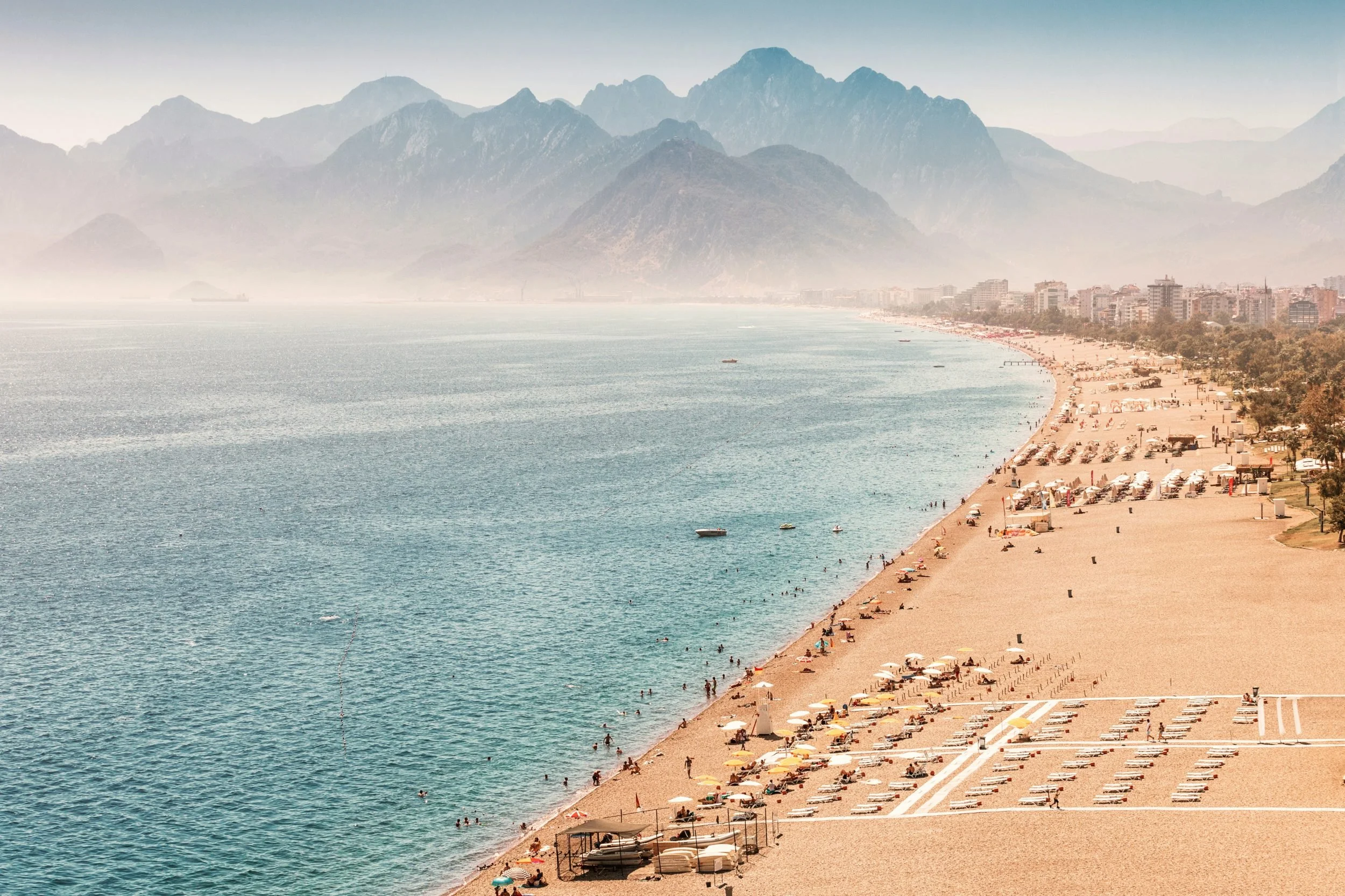 A beach with umbrellas and lounge chairs, shoreline with people swimming, and a city with mountains in the background.