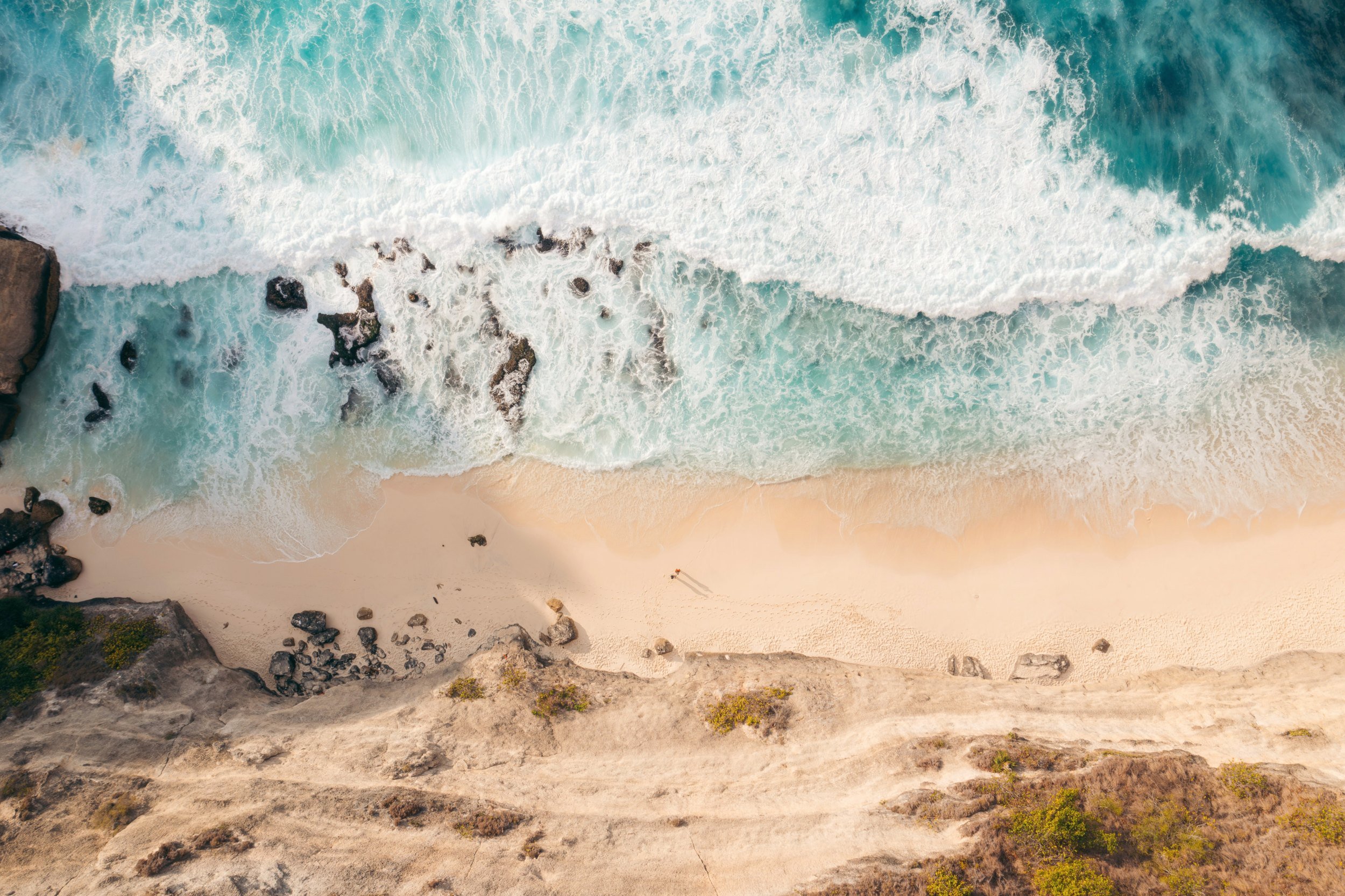 Aerial view of a sandy beach with turquoise ocean waves crashing against rocks and shoreline, with some vegetation on the cliff edge.