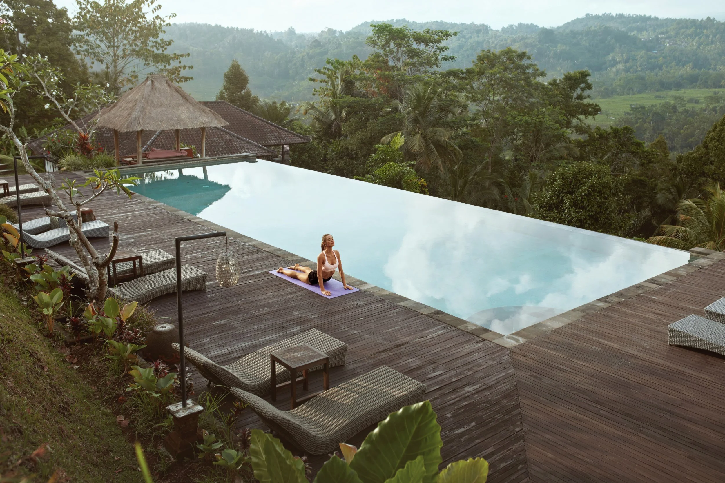 A person practicing yoga by a infinity-edge swimming pool on a wooden deck with a lush green forest and mountains in the background.
