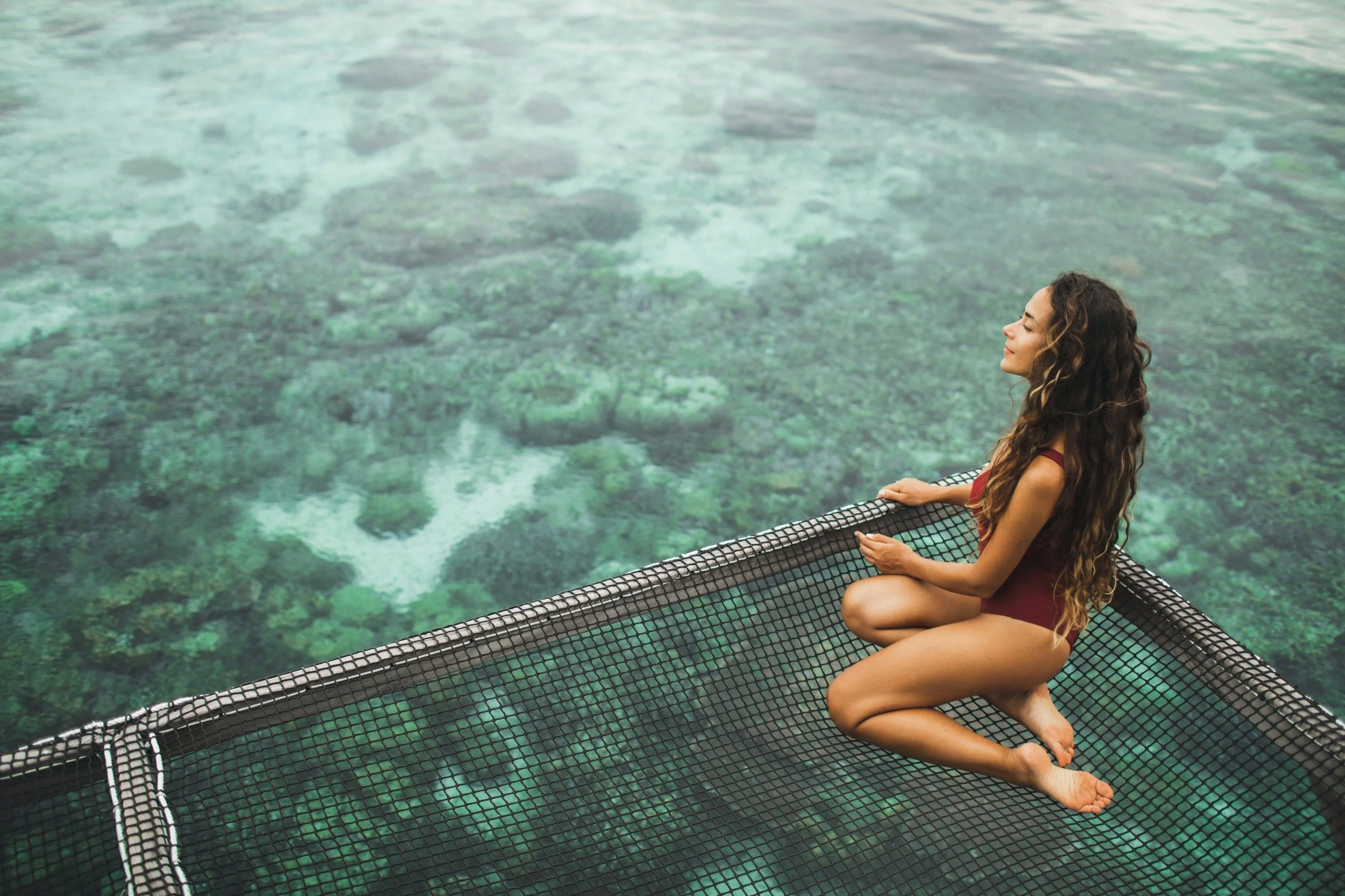 Woman meditating on a netted platform above clear turquoise water with visible rocks and coral underneath.