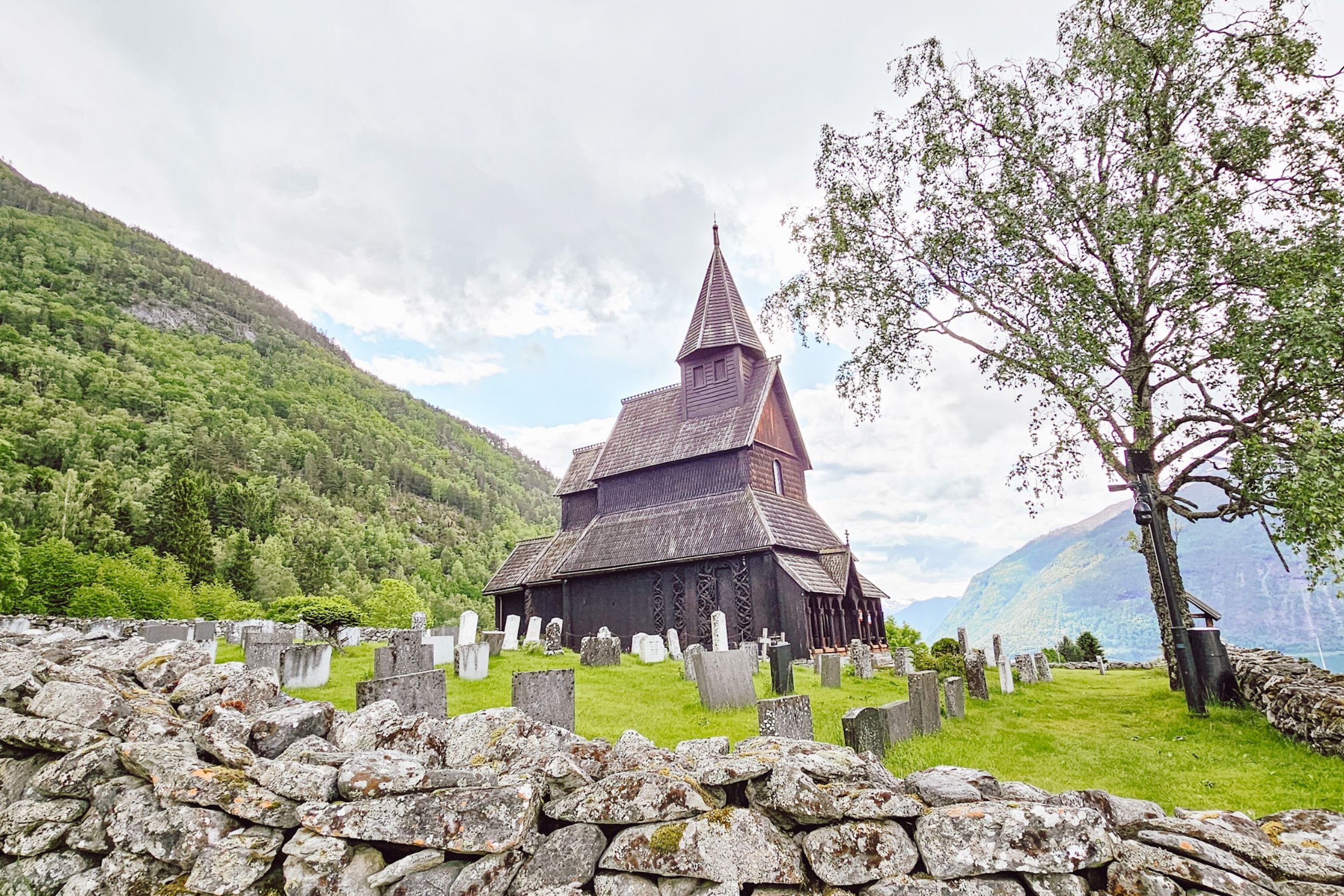 Wooden stave church in the middle of a field surrounded by tombstones and hills covered with trees.