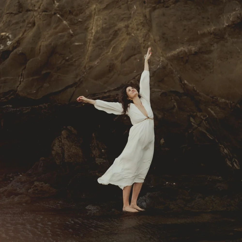 Woman in a white dress standing on the shore in front of a rock with both arms reaching towards the sky