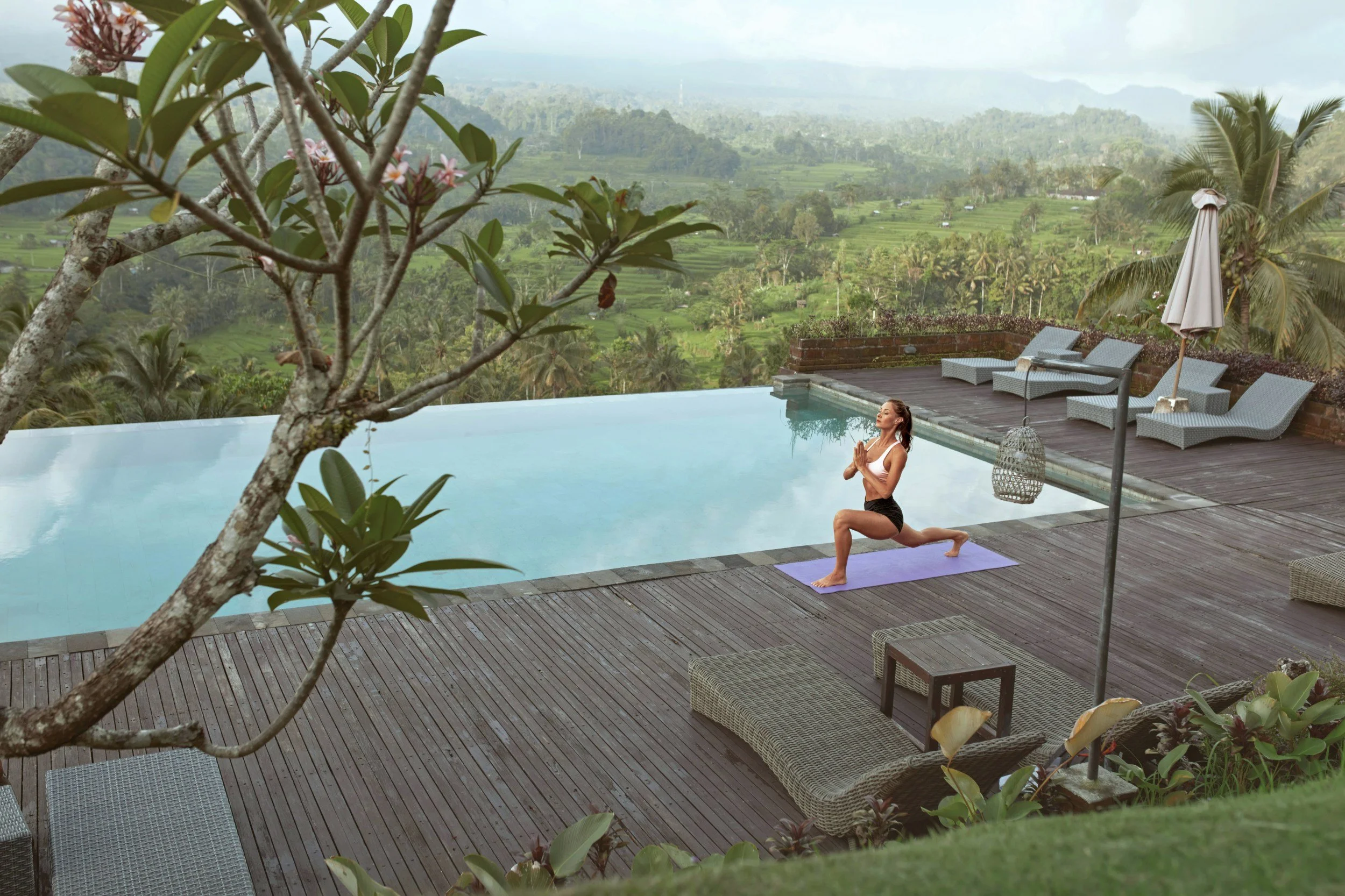 A woman practicing yoga on a purple mat next to a pool on a wooden deck with lush green landscape in the background.