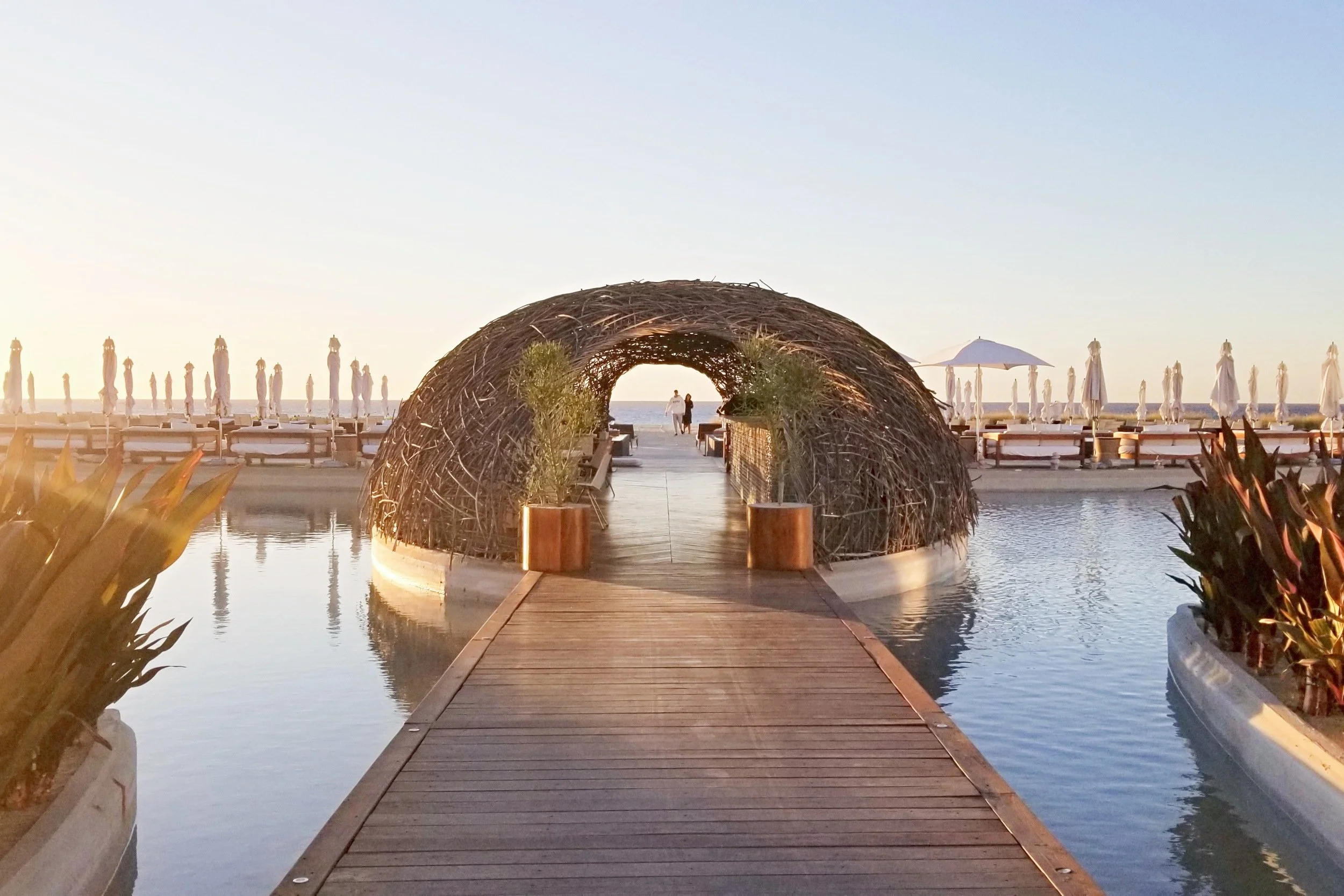 A wooden walkway leading to a large, round woven nest-like structure at the beach, with lounge chairs and umbrellas in the background, and the sunset sky overhead.