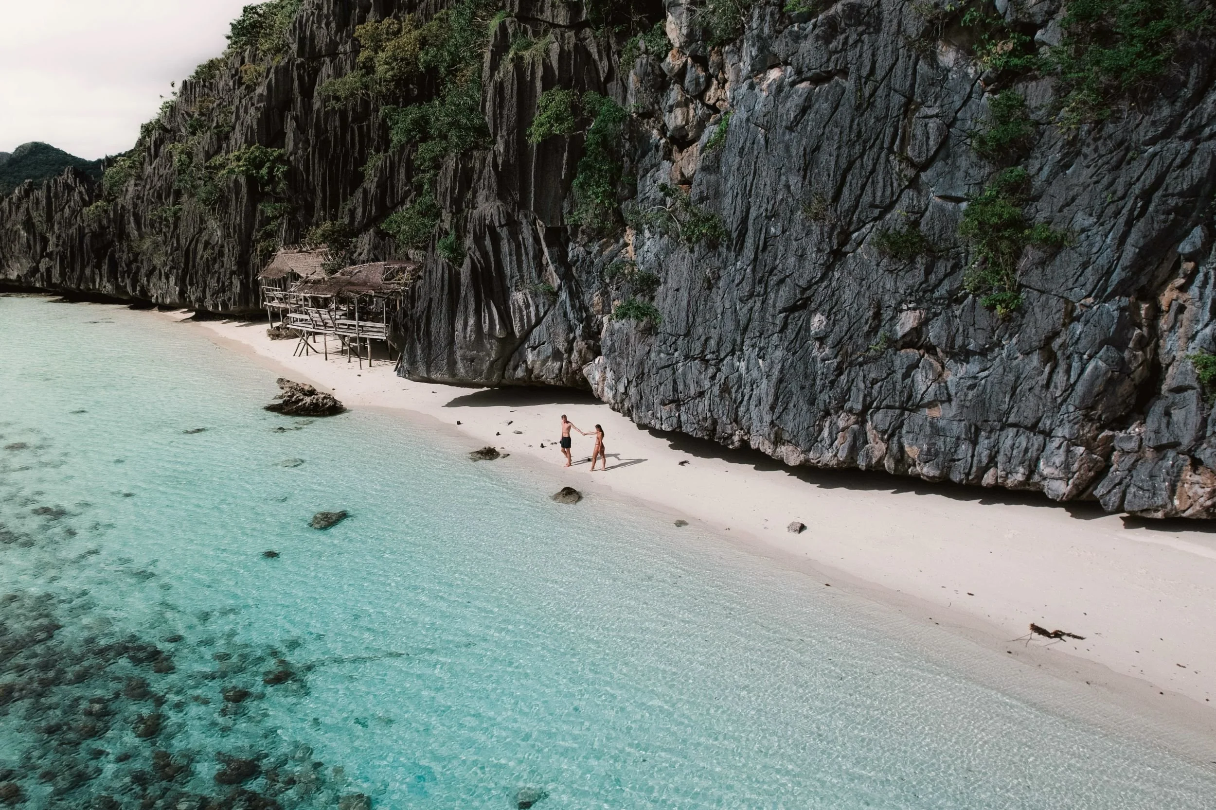 People walking on a sandy beach with clear turquoise water, large rocks, and tall dark gray cliffs with green vegetation.