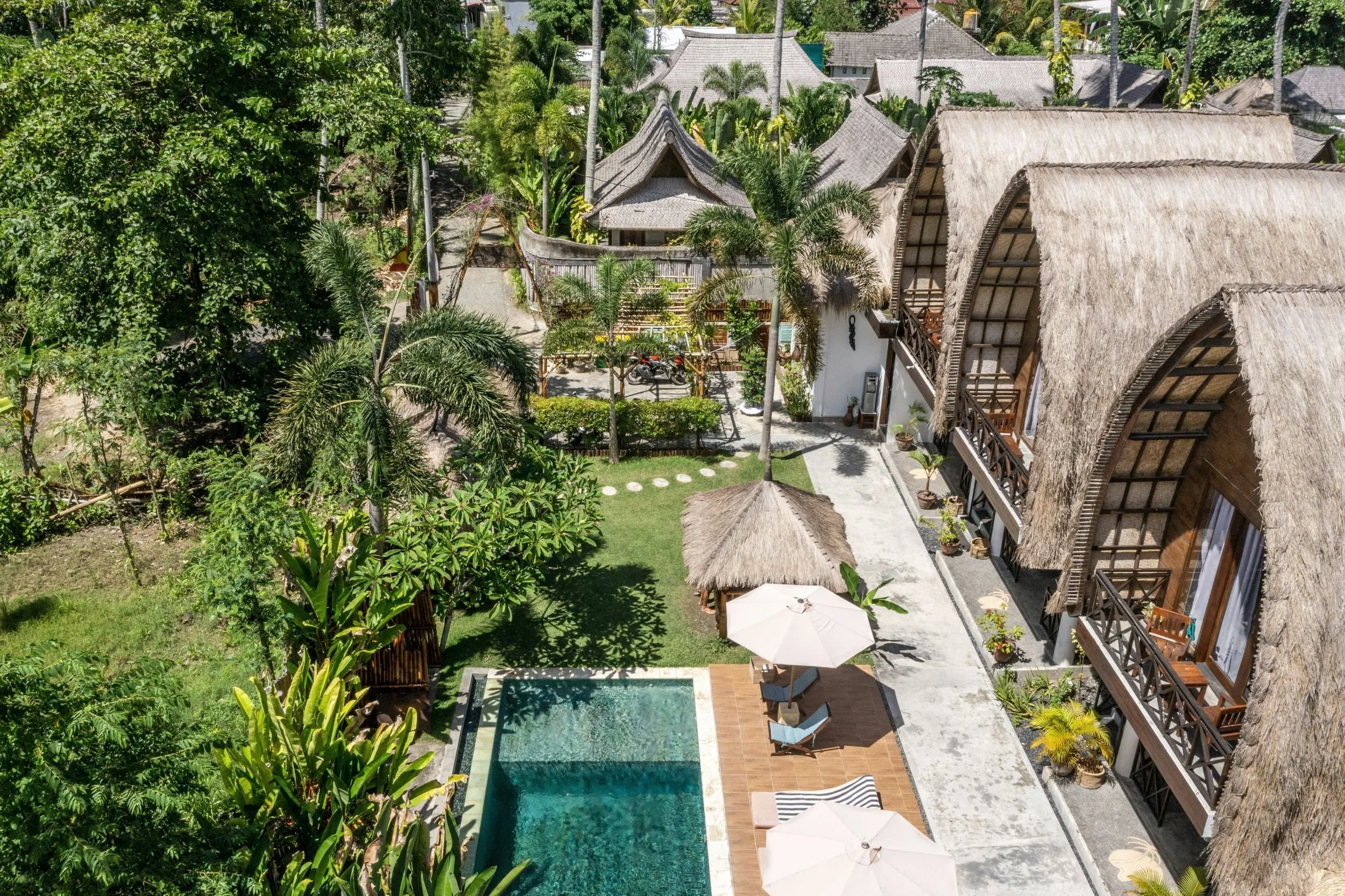 Aerial view of a resort with thatched roof buildings, a swimming pool, a grassy yard with palm trees, and outdoor seating with umbrellas.