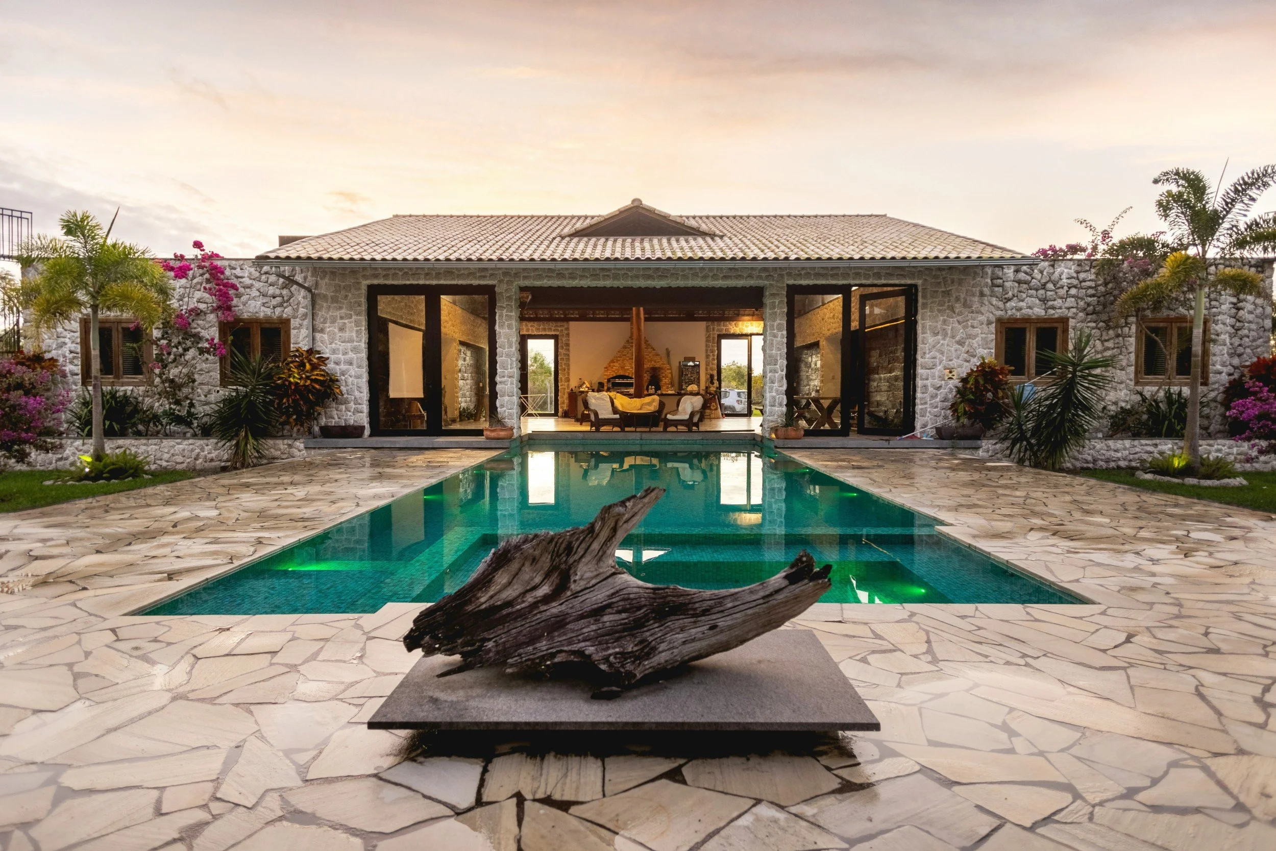 Luxury house with a stone exterior, swimming pool, and tropical plants at sunset, interior visible through large glass doors.