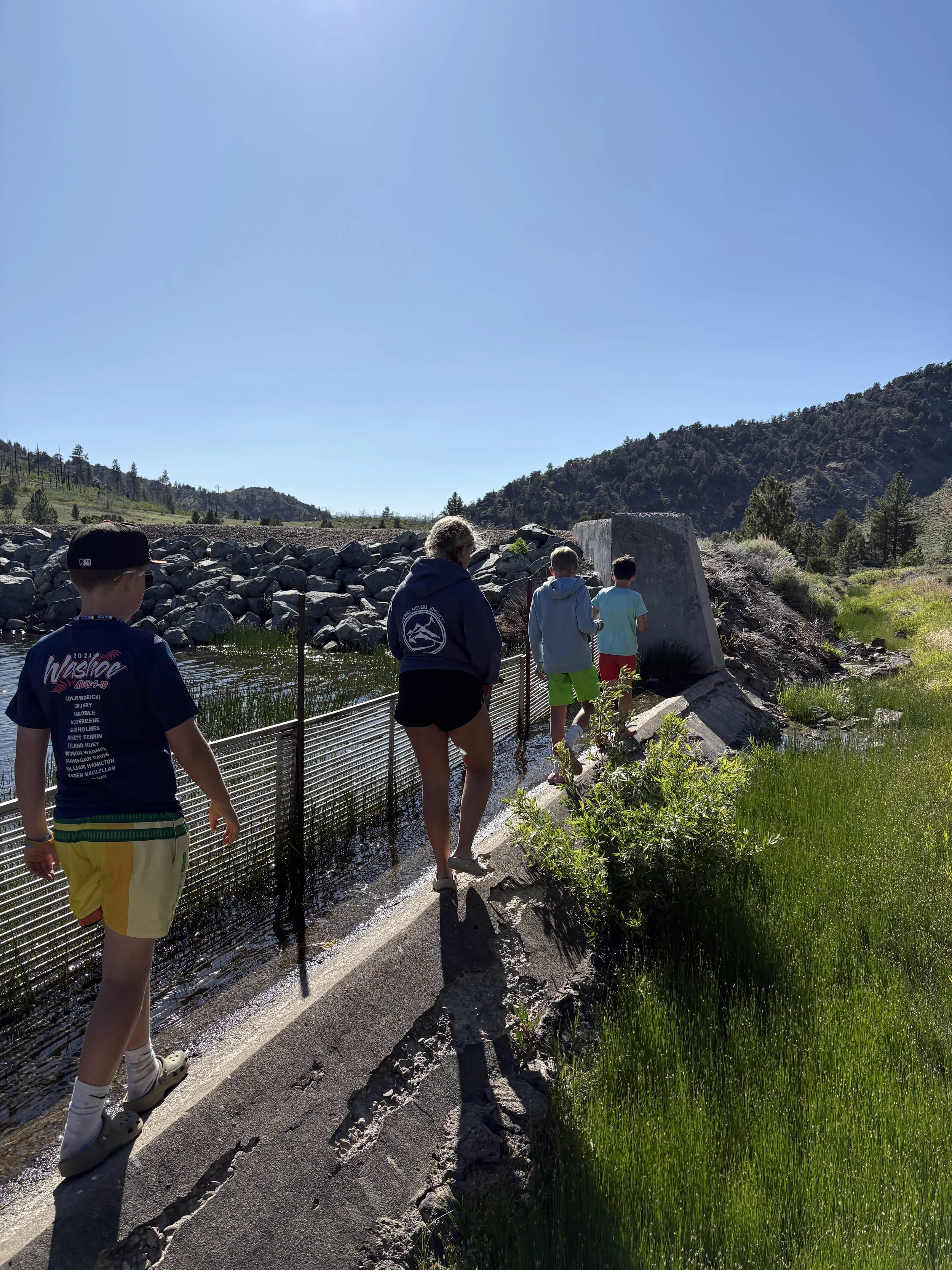 People walking along a narrow concrete path beside a pond with rocks on the opposite side, in a mountainous outdoor setting on a sunny day.