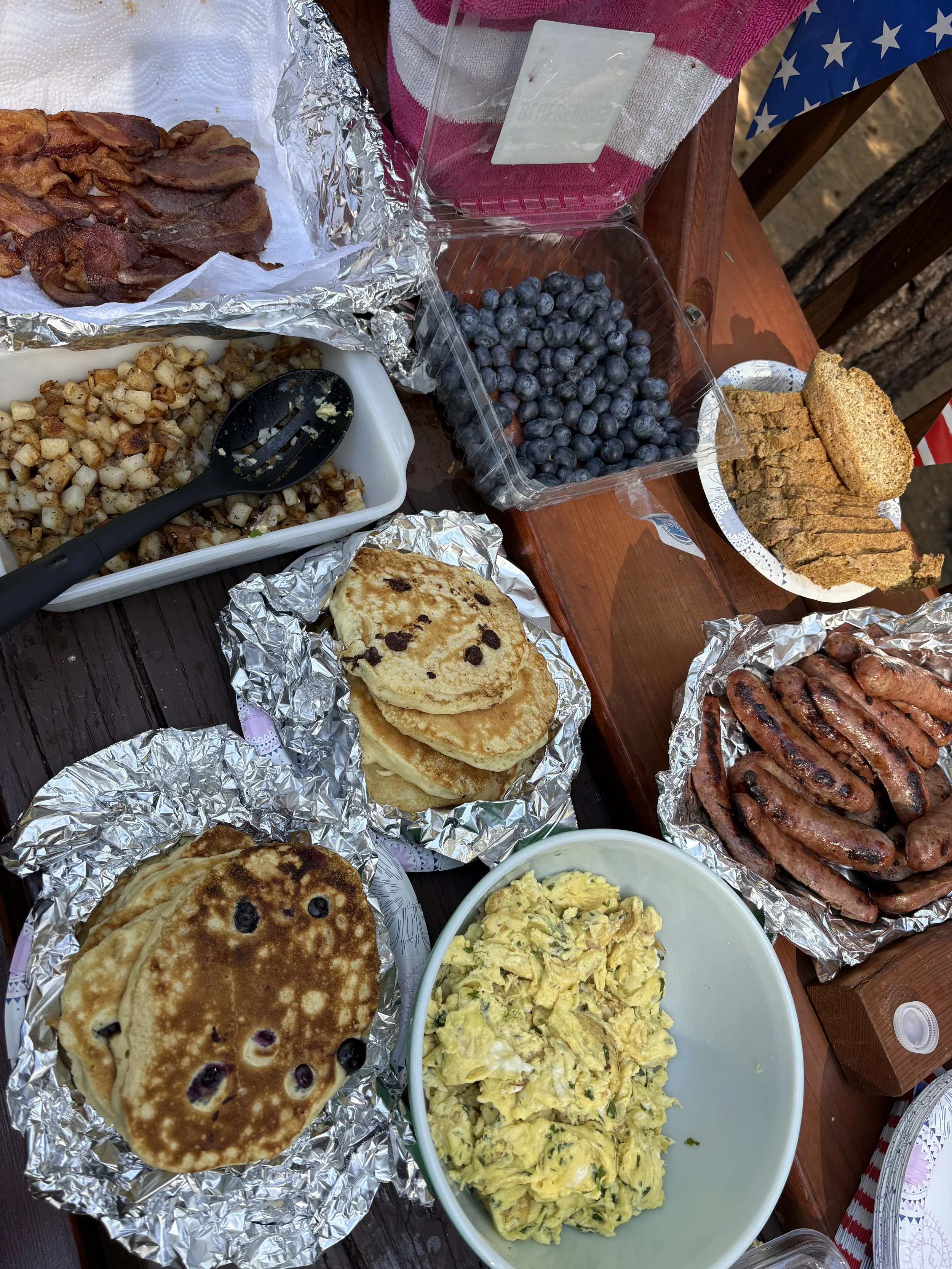 A variety of breakfast foods on a picnic table, including bacon, blueberry pancakes, scrambled eggs, grilled sausages, and cornbread, with a container of blueberries and a pink drink in a clear cup.