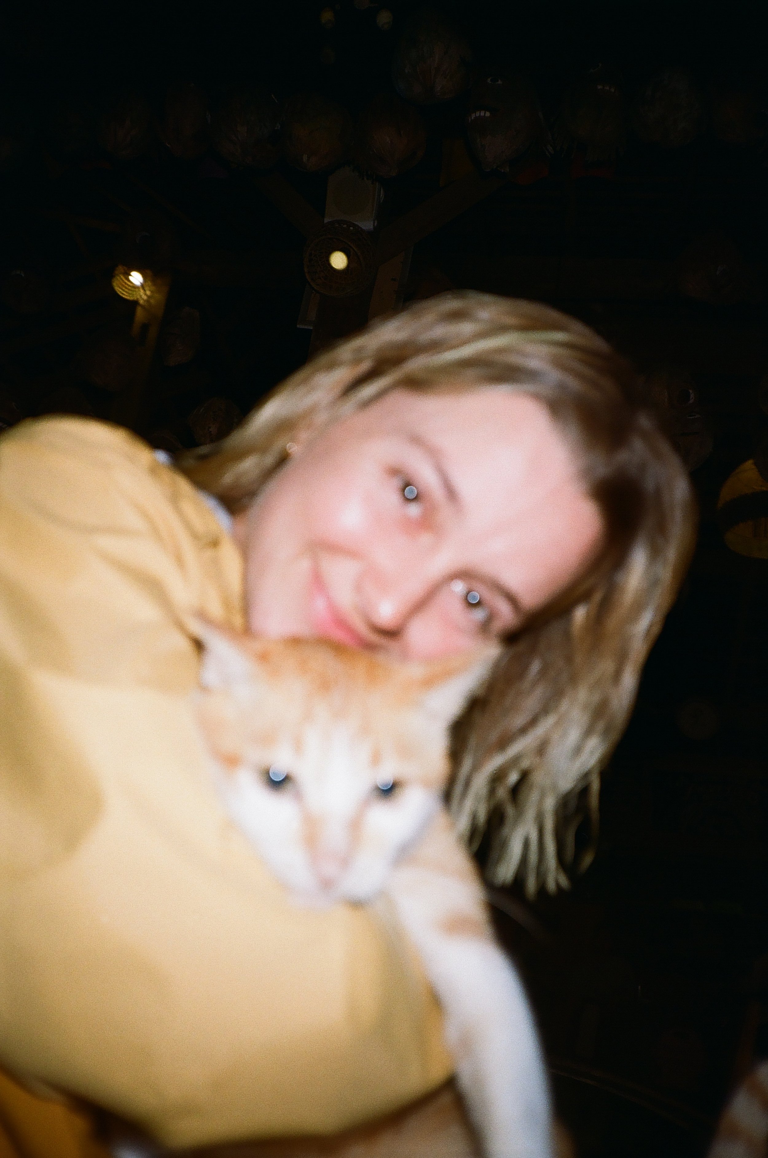 A young woman with blonde hair holding a light-colored cat close to her face, both looking towards the camera, in a dimly lit environment.