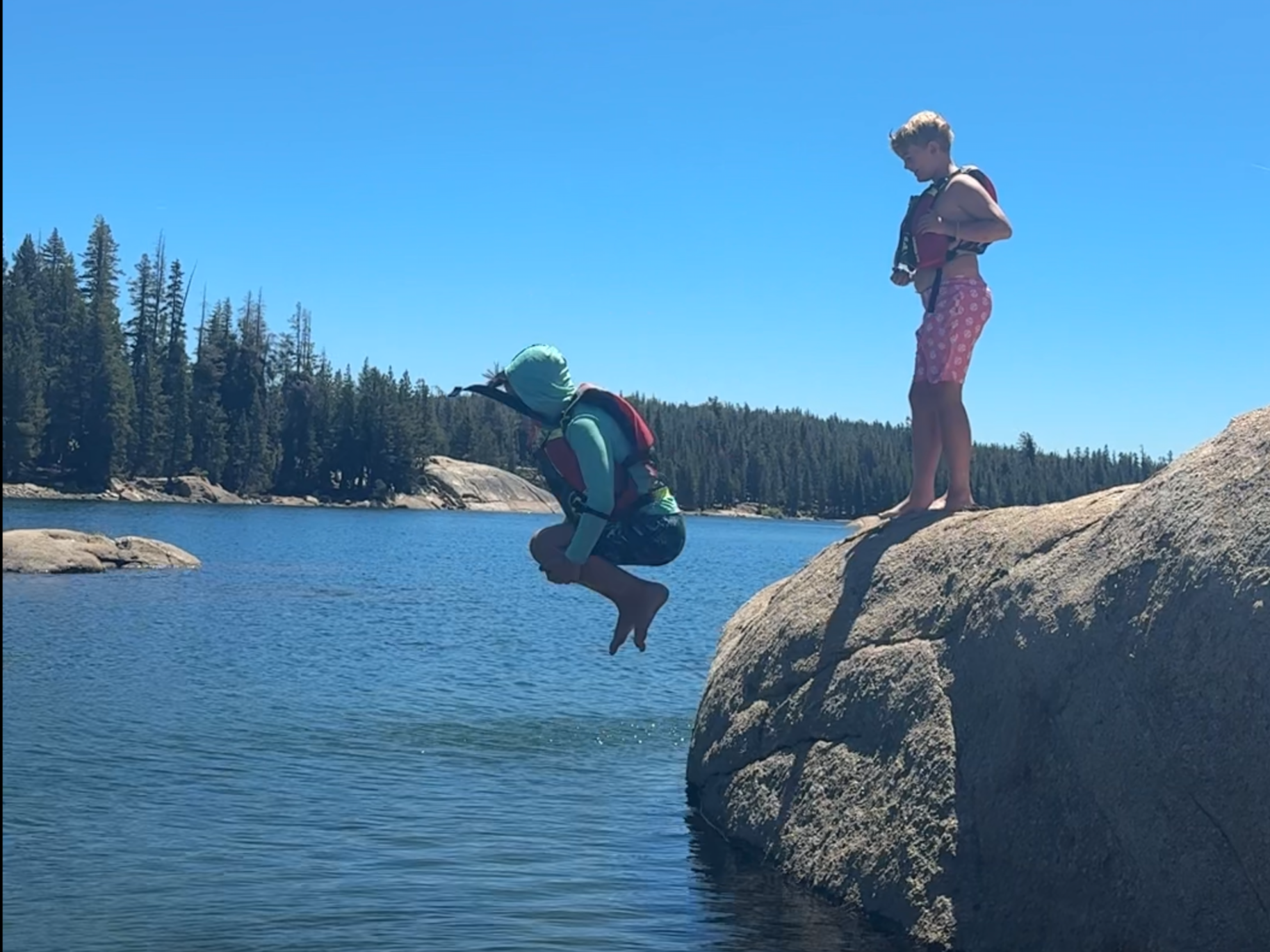 A person in a hoodie and life jacket is in mid-air, jumping from a large rock into a lake. Another person, in a swimsuit and also with a life jacket, stands on the rock watching. The background features a pine forest under a clear blue sky.