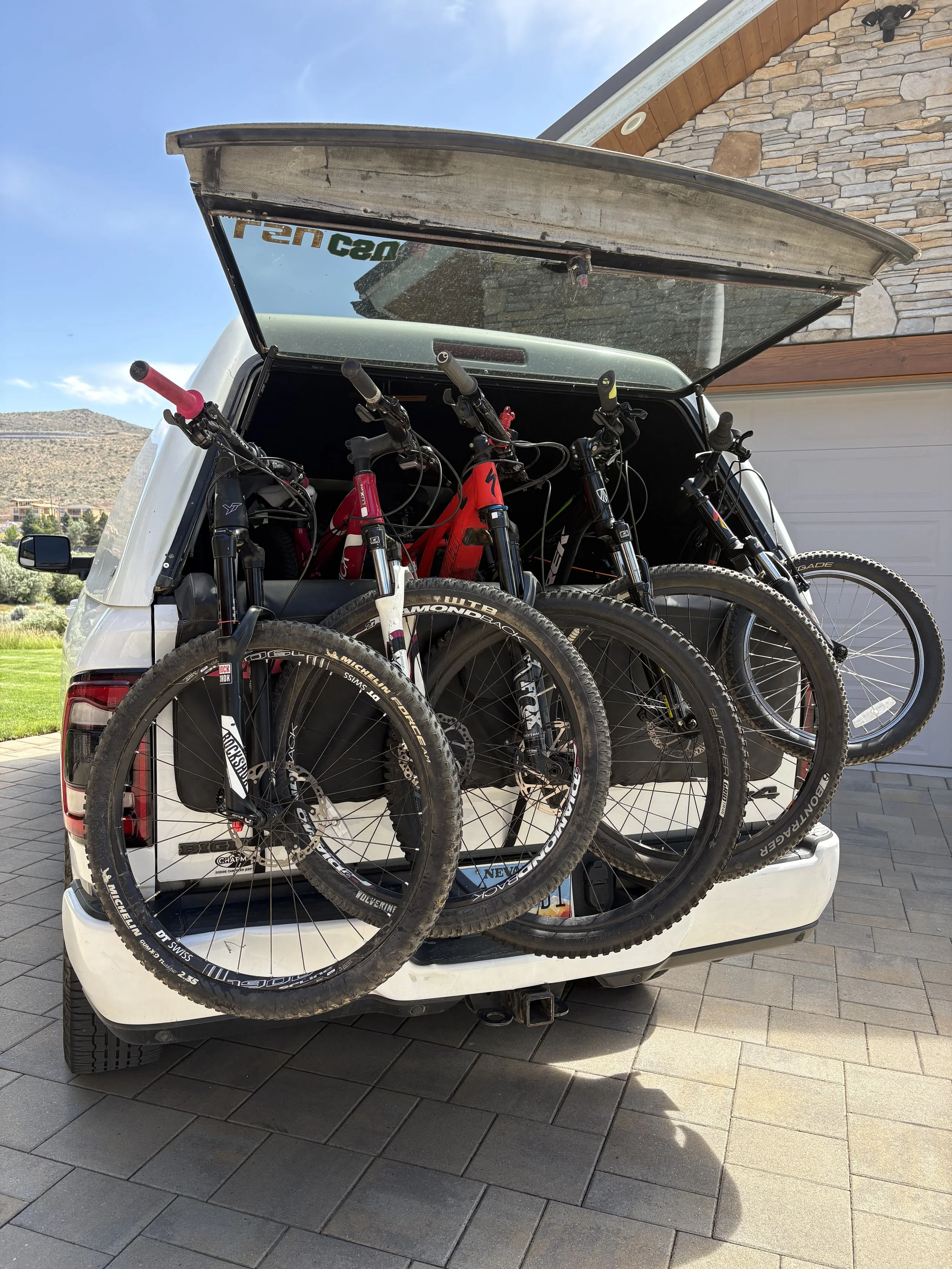 Open tailgate of a white SUV with four mountain bikes mounted inside in a parking lot with a house, a green lawn, and mountains in the background.