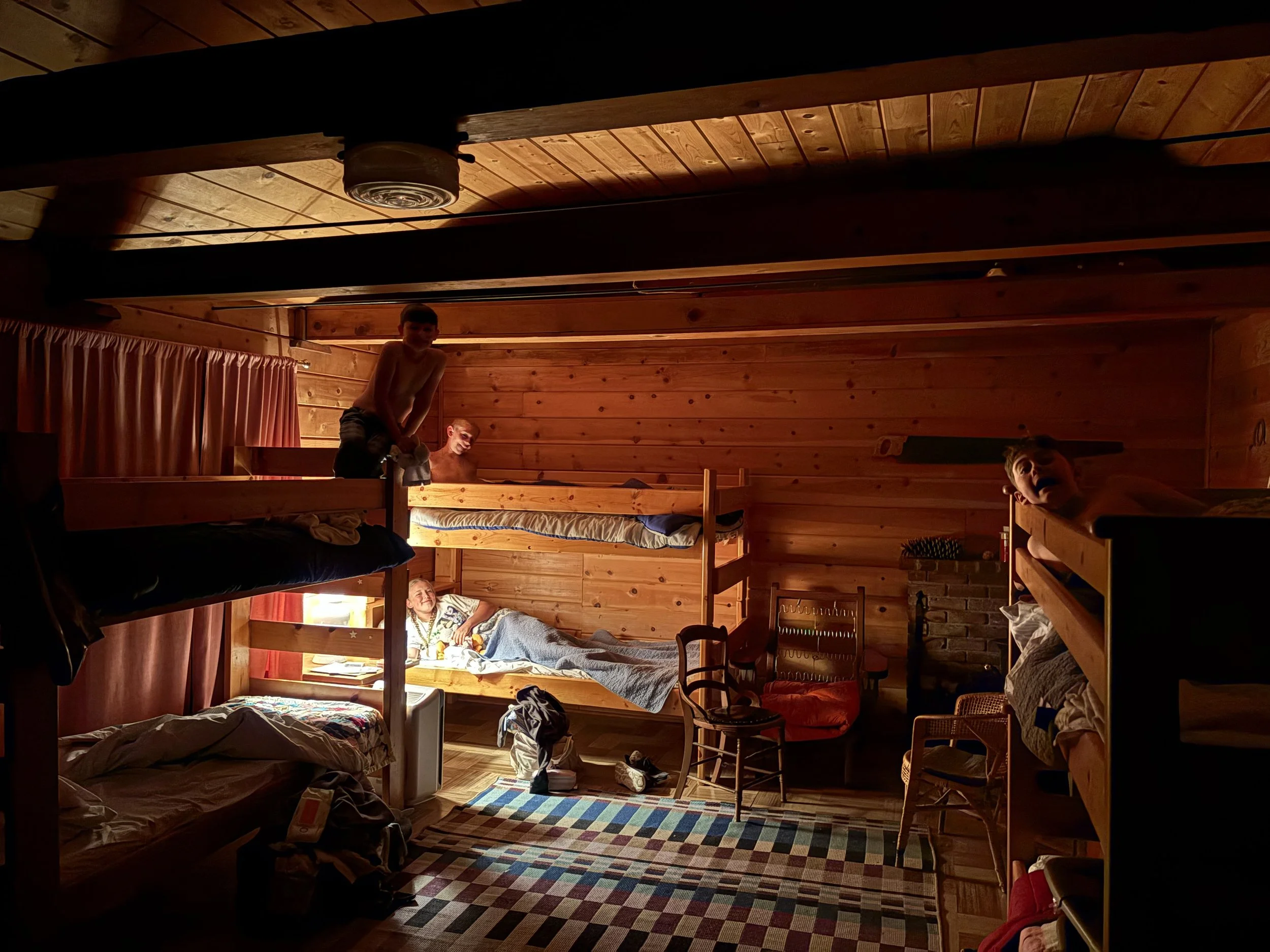 A wooden-paneled room with bunks, with four children visible, some lying or sitting on beds, one standing, and one peeking from a bed. The room has a rustic, cozy appearance with a checkerboard rug and chairs.